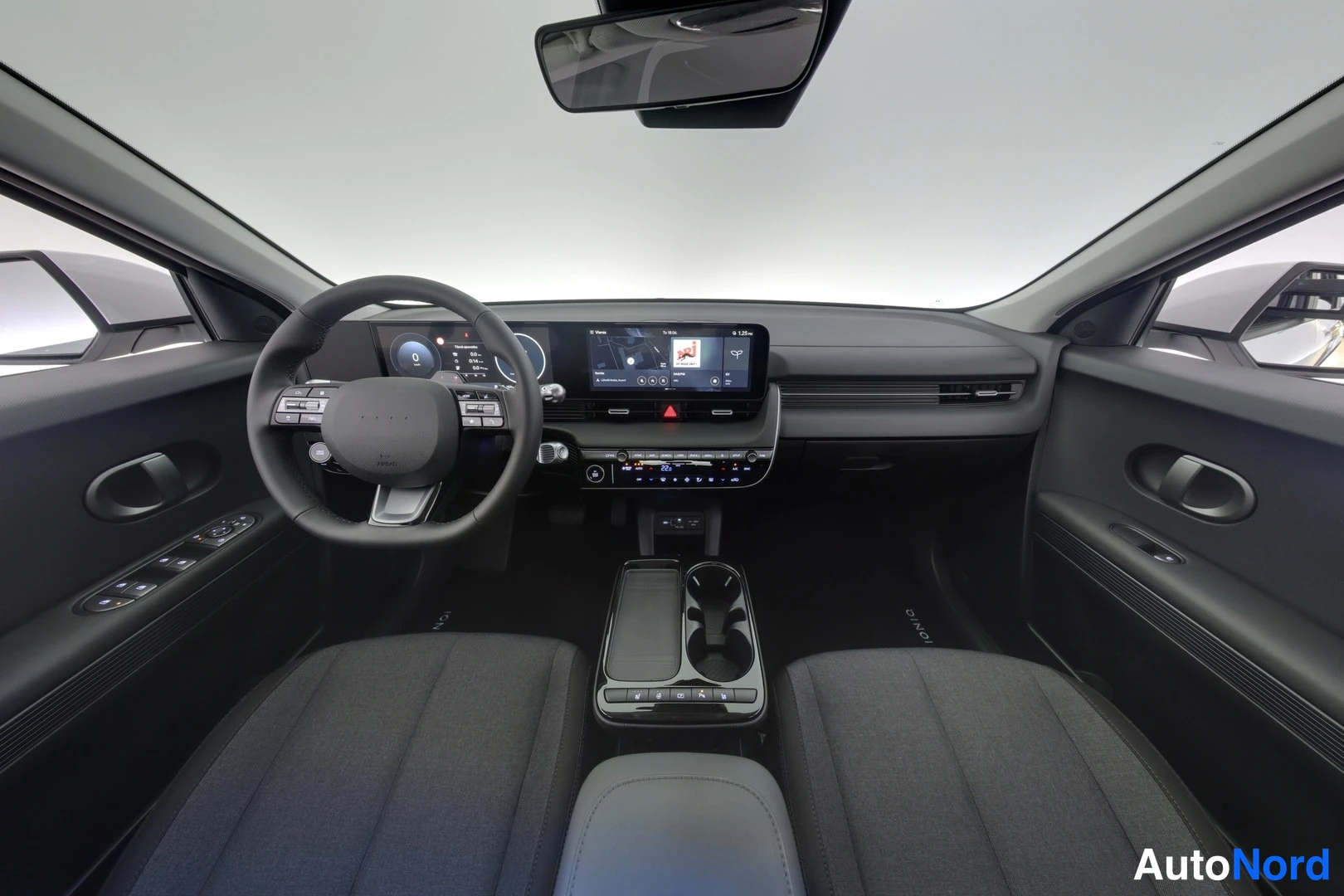 Muu: Hyundai IONIQ 5:n ohjaamo etäältä kuvattuna: ohjaustehostin, digitaalinen kojelauta, keskikonsolin kosketusnäyttö ja matala keskitunneli. Istuimet tummaa teksti