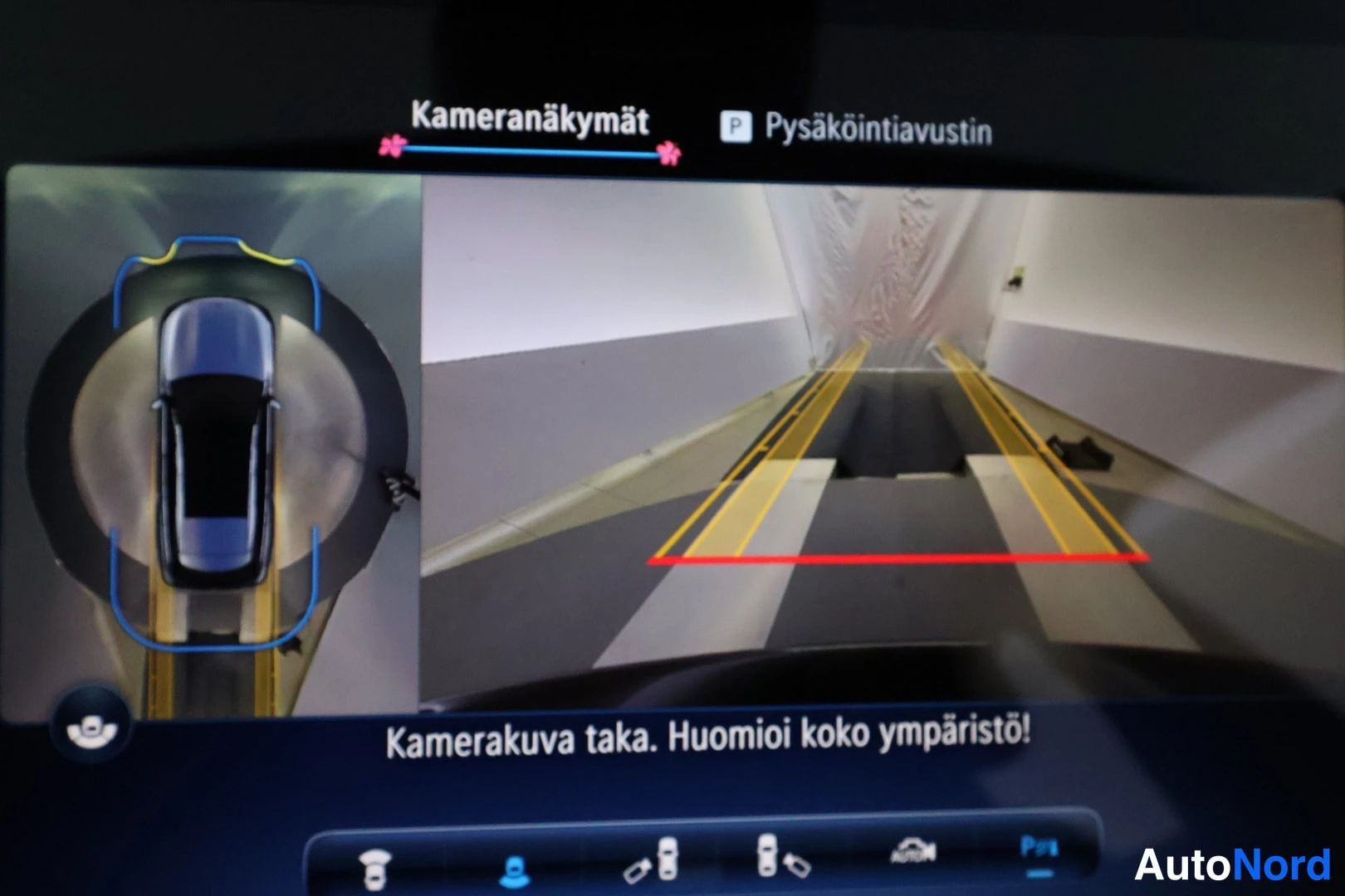 Muu: Näytön kameraäkymä, jossa vasemmalla auton ympärysnäkymä ja oikealla peruutuskameran kuva tekstillä 'Kamerakuva taka. Huomioi koko ympäristö!'.
