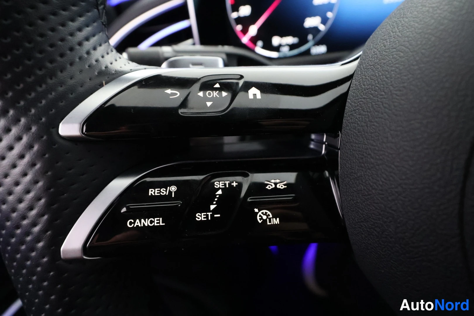 Yksityiskohta: Lähikuva Mercedes-Benzin ohjauspyörän vasemmasta pikelärästä: napit RES/|, CANCEL, SET +, SET -, LIM, kulmavalot sekä OK-nuolinappi ja kotinappi; taustalla tumm