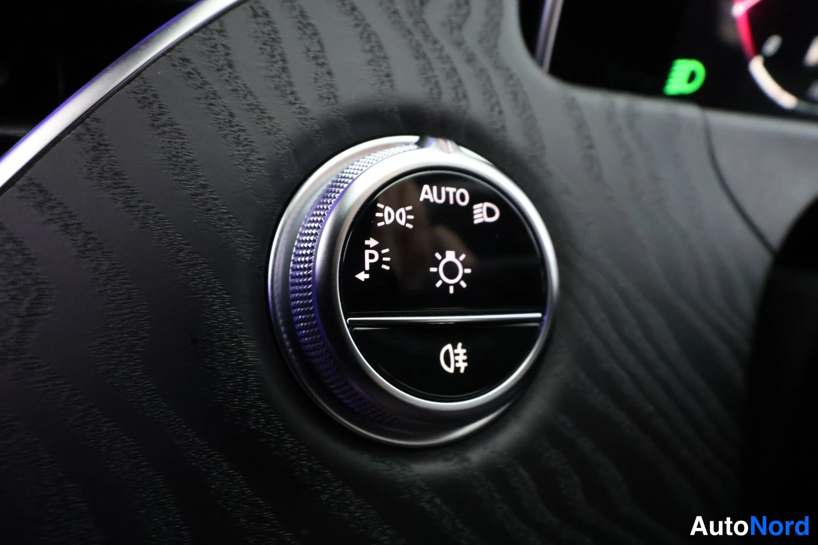 Yksityiskohta: Lähikuva Mercedes-Benzin kojelaudan pyöreästä valokytkimestä: nupissa AUTO, sumuvalot, pysäköintivalot, valonsäätö ja sumutakselin kuvake; taustalla vihreä D-me