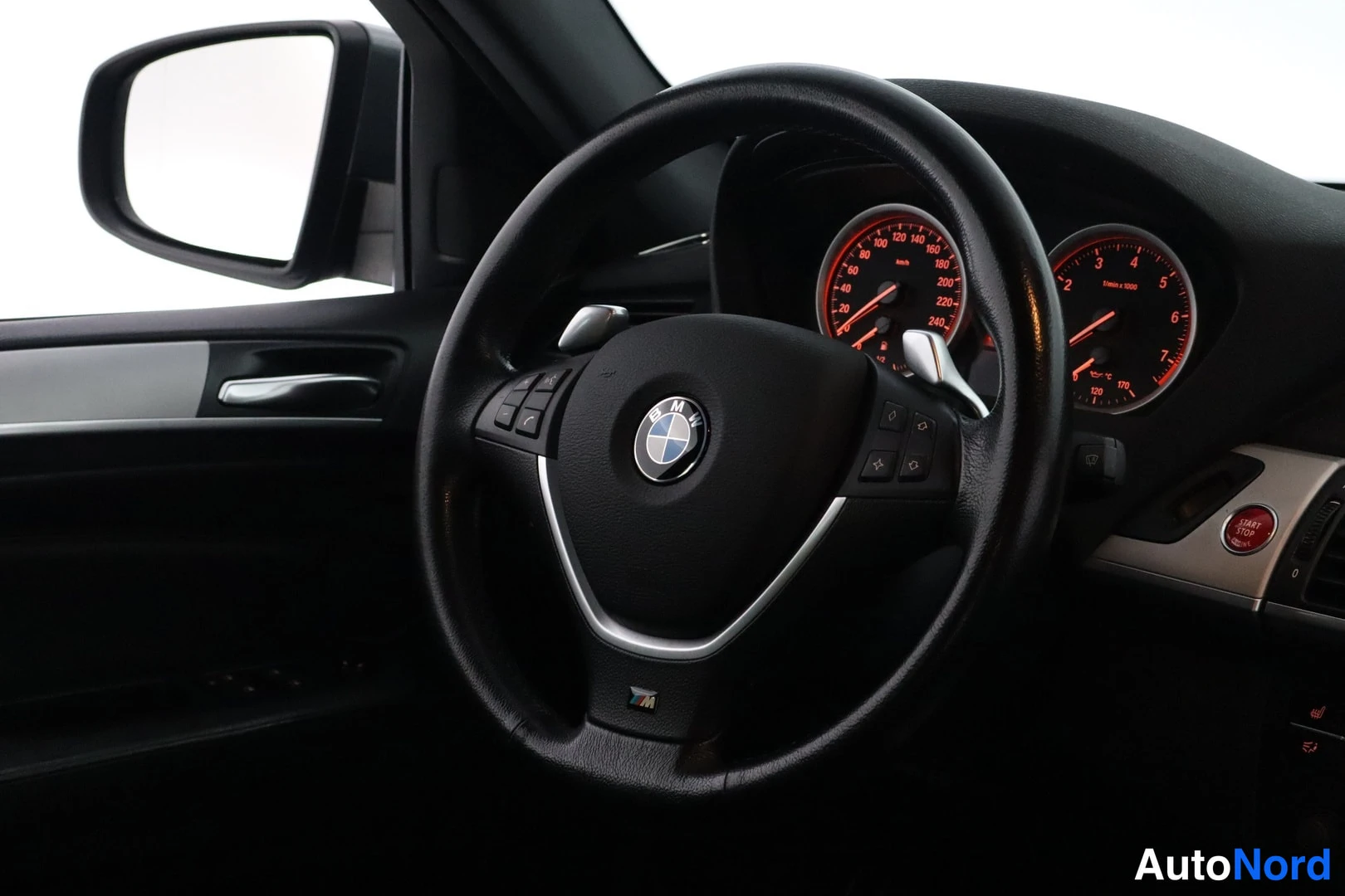 Takaa: BMW-ohjauspyörä ja punaisella valaistu kojeisto kuvattuna läheltä, vasemmalla sivupeili.