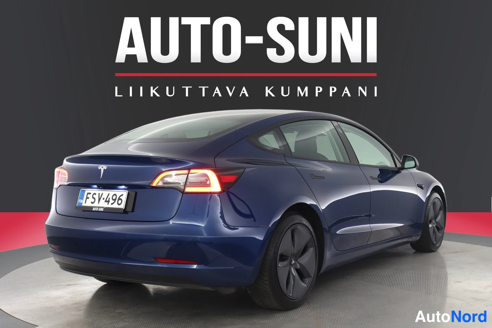 Takaviisto: Tummansininen sedan takaa hieman sivulta, takavalot päällä, rekisterikilpi.