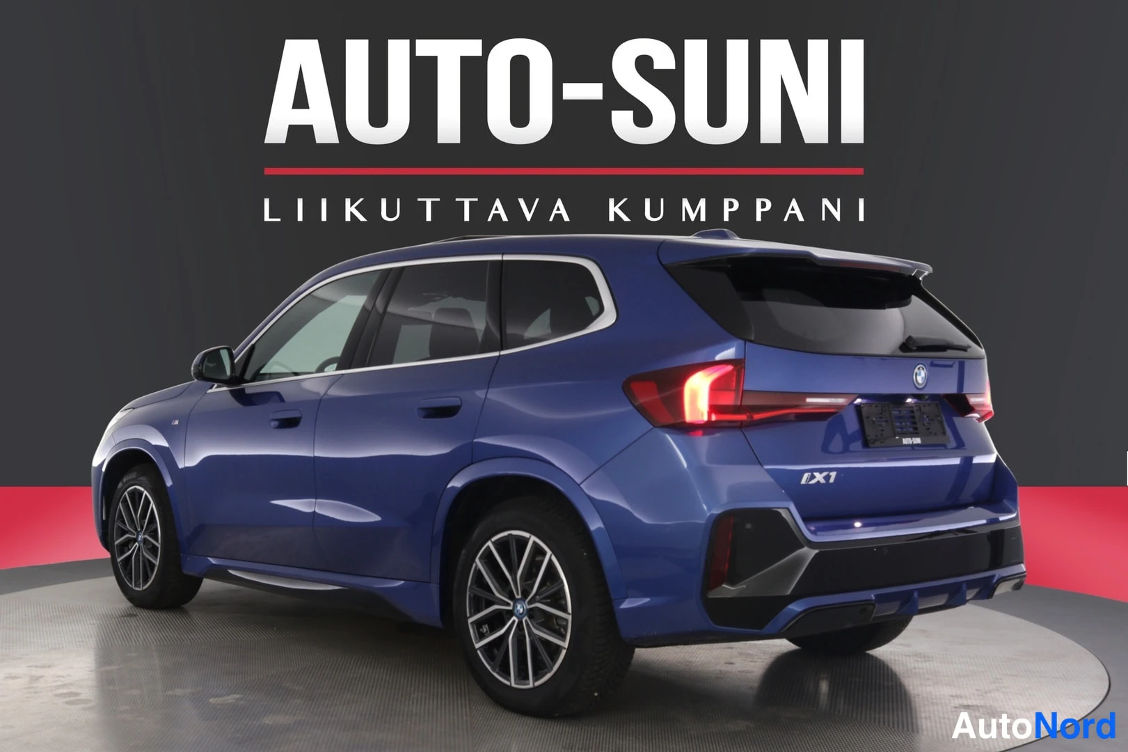 Takaviisto: Sininen BMW iX1 toisesta takakulmasta, takavalo sytytettynä.
