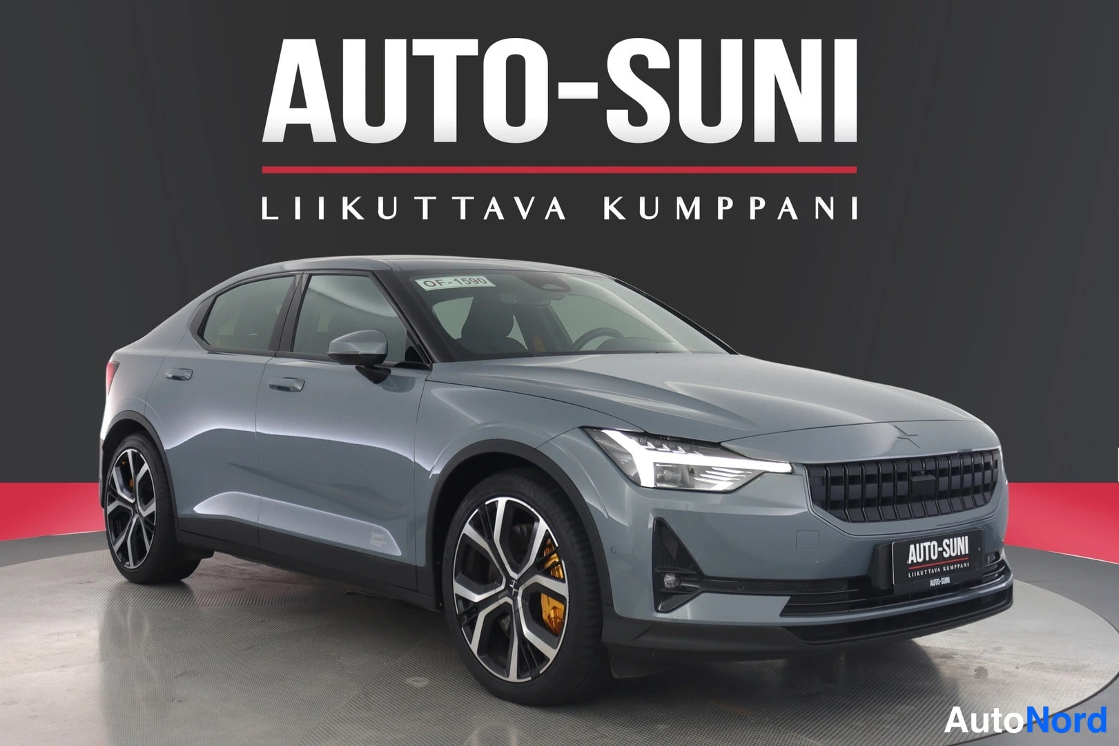 Etuviisto, nokka oikealle: Sinertävänharmaa henkilöauto kuvattuna etukolmanneksesta näyttelylavalla, AUTO-SUNI-mainos taustalla.