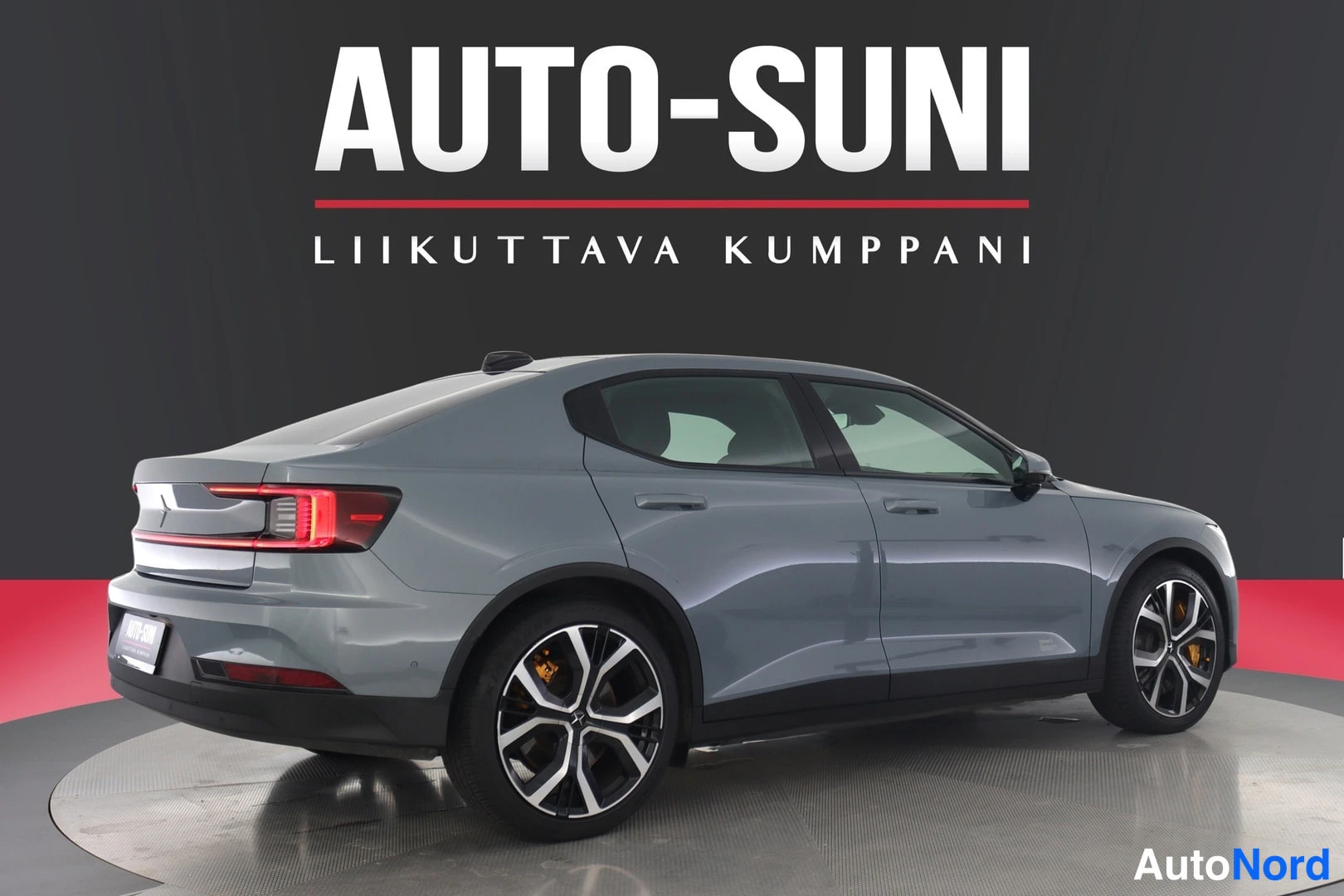 Takaviisto: Sinertävänharmaan henkilöauton takakolmanneksesta otettu kuva näyttelylavalla, AUTO-SUNI-mainos taustalla.