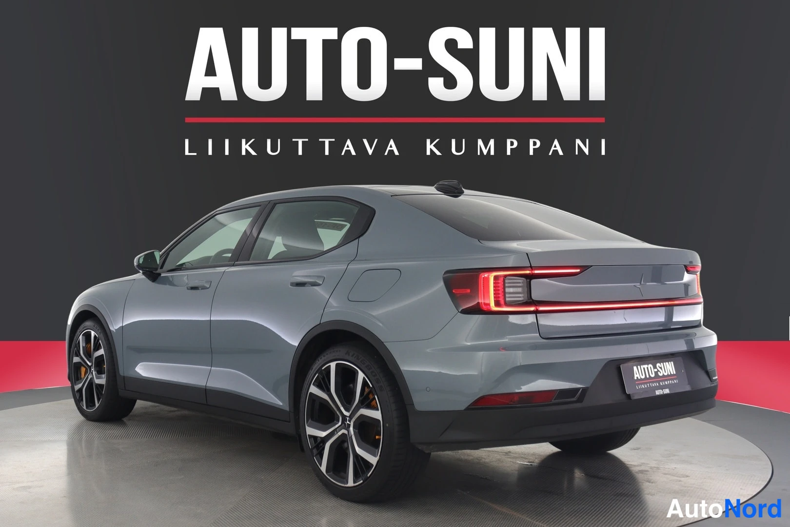 Takaviisto: Sinertävänharmaa henkilöauto takakolmanneksesta näyttelylavalla, AUTO-SUNI-kyltti perällä.