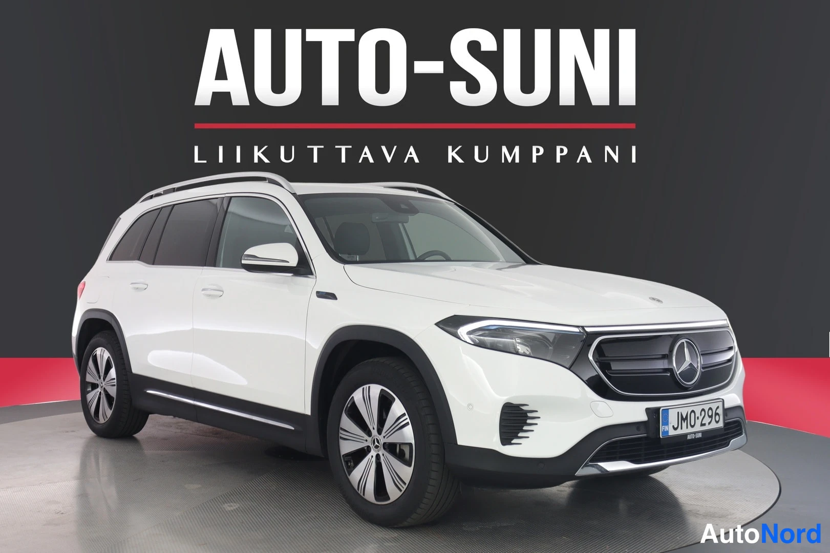 Etuviisto, nokka oikealle: Valkoinen Mercedes-Benz EQB -tyyppinen farmarimallinen auto kuvattu kolmelinjaisesta etukulmasta oikealta; kattokaiteet, rekisterikilpi, AUTO-SUNI-tausta
