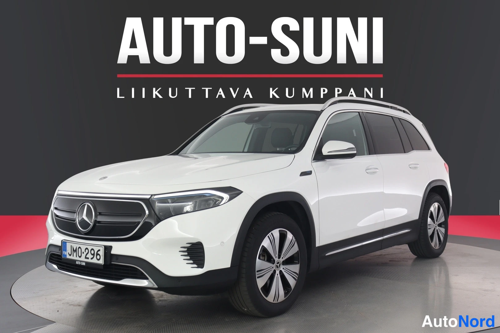 Etuviisto, nokka vasemmalle: Valkoinen Mercedes-Benz EQB etukulmasta, rekisterikilpi JM0-296, taustana AUTO-SUNI-mainosnurkisto.