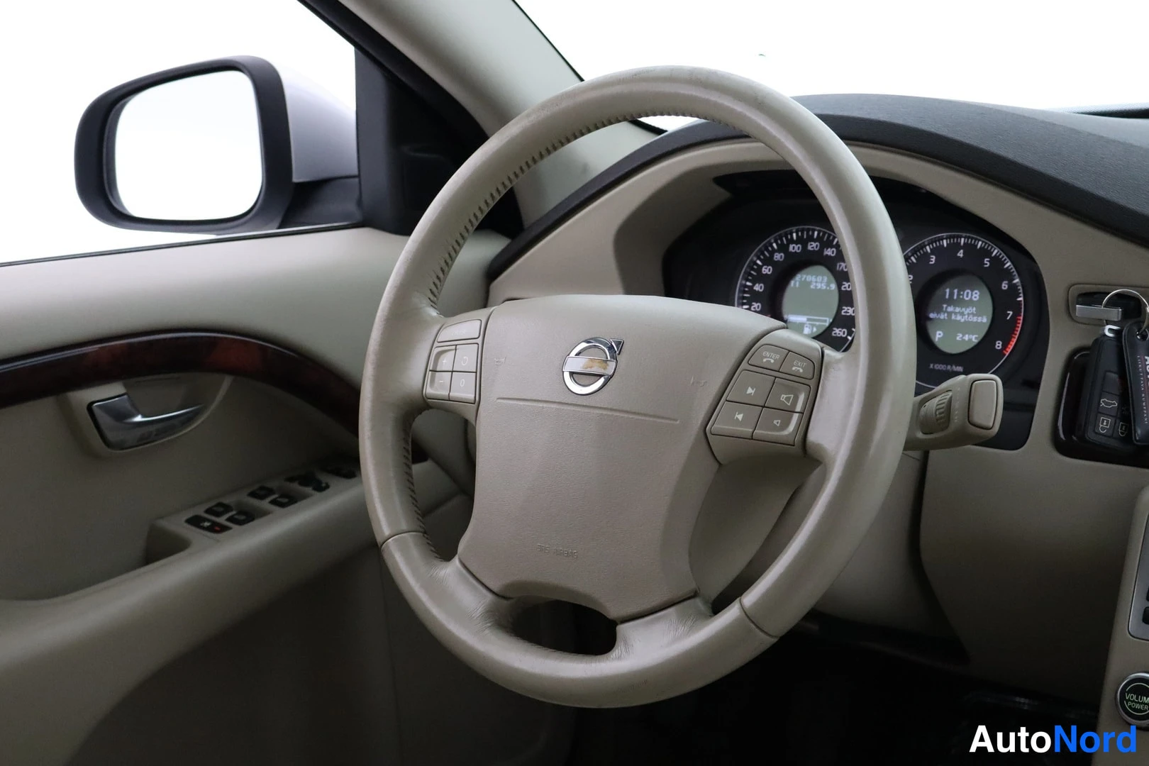 Muu: Volvo-ohjauspyörä ja kojelauta, mittaristo näkyy, vaalea beige sisustus, taustapeili vasemmalla.