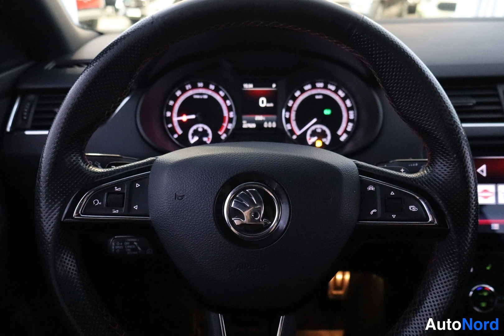 Muu: Ohjauspyörä Skoda-merkillä ja monitoiminäppäimillä, mittaristo taustalla.