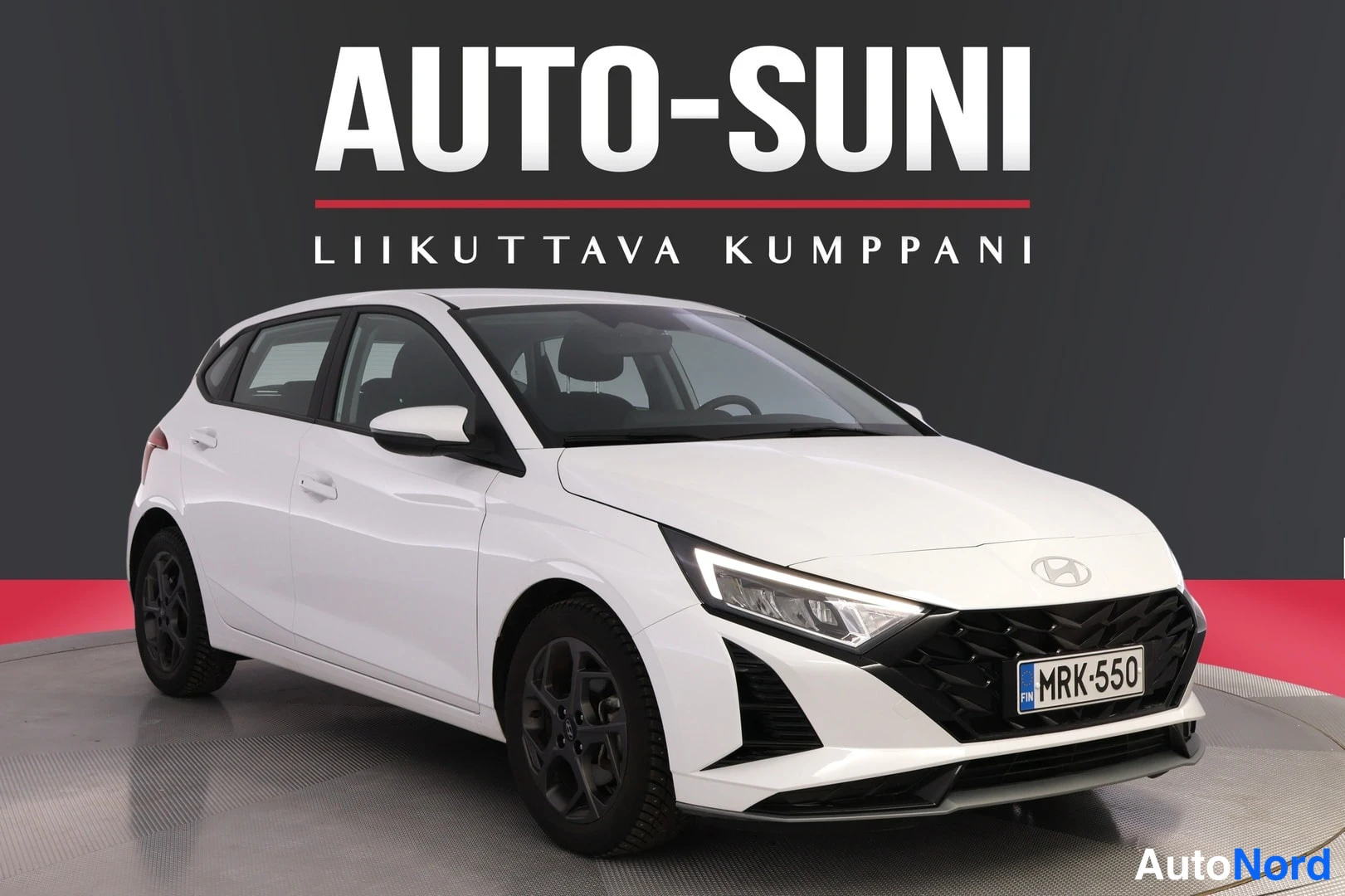 Etuviisto, nokka oikealle: Valkoinen Hyundai i20 -henkilöauto kuvattu etuviistosta oikealle kääntyneellä keulalla, rekisterikilpi, harmaa studiosäädö taustalla.