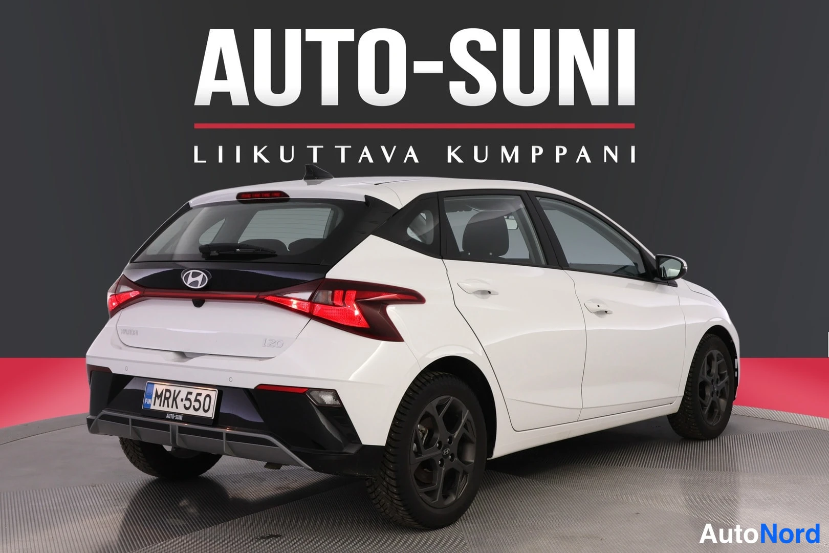 Takaviisto: Valkoinen Hyundai i20 -henkilöauto takaviistosta, punaiset takavalot ja rekisterikilpi näkyvissä.