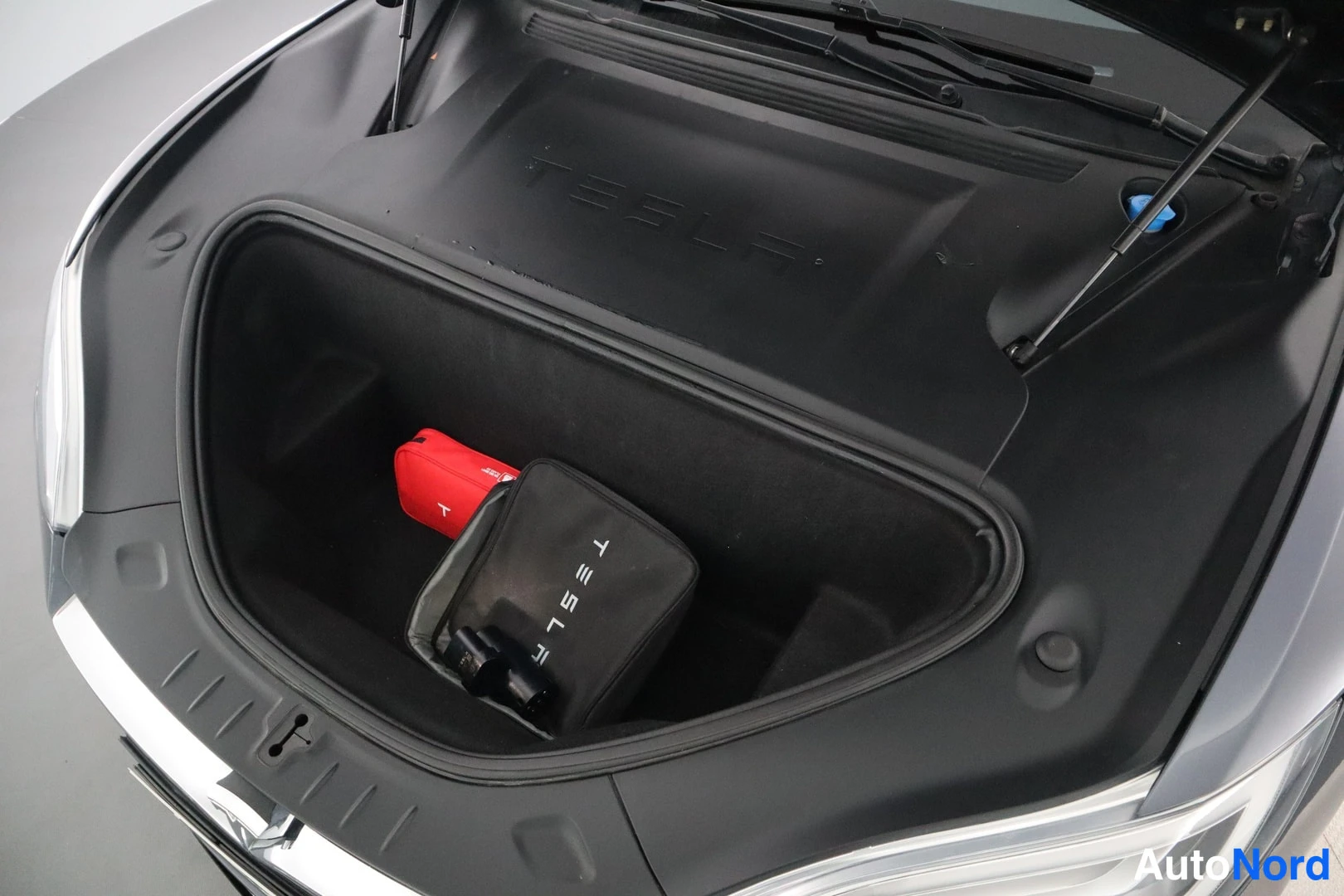 Muu: Auton etutavaratila avattuna; sisällä punainen laukku ja Tesla-merkitty musta laukku.