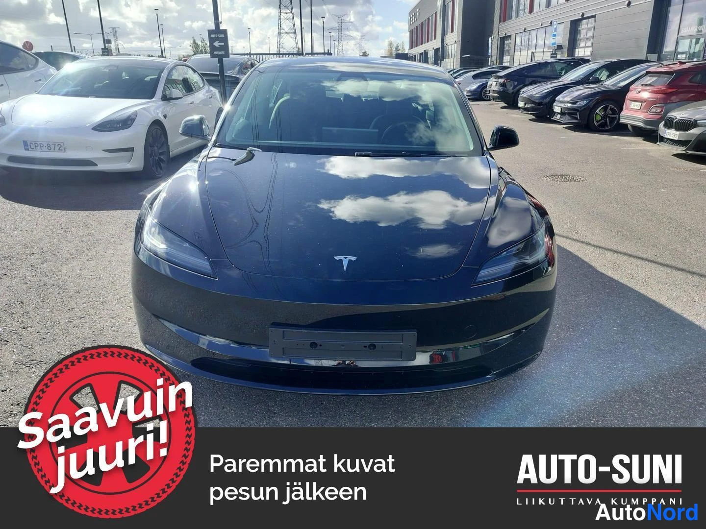Edestä: Mustan Tesla Model 3 -henkilöauton etunäkyä sisäänajopaikalla, muut autoja taustalla.