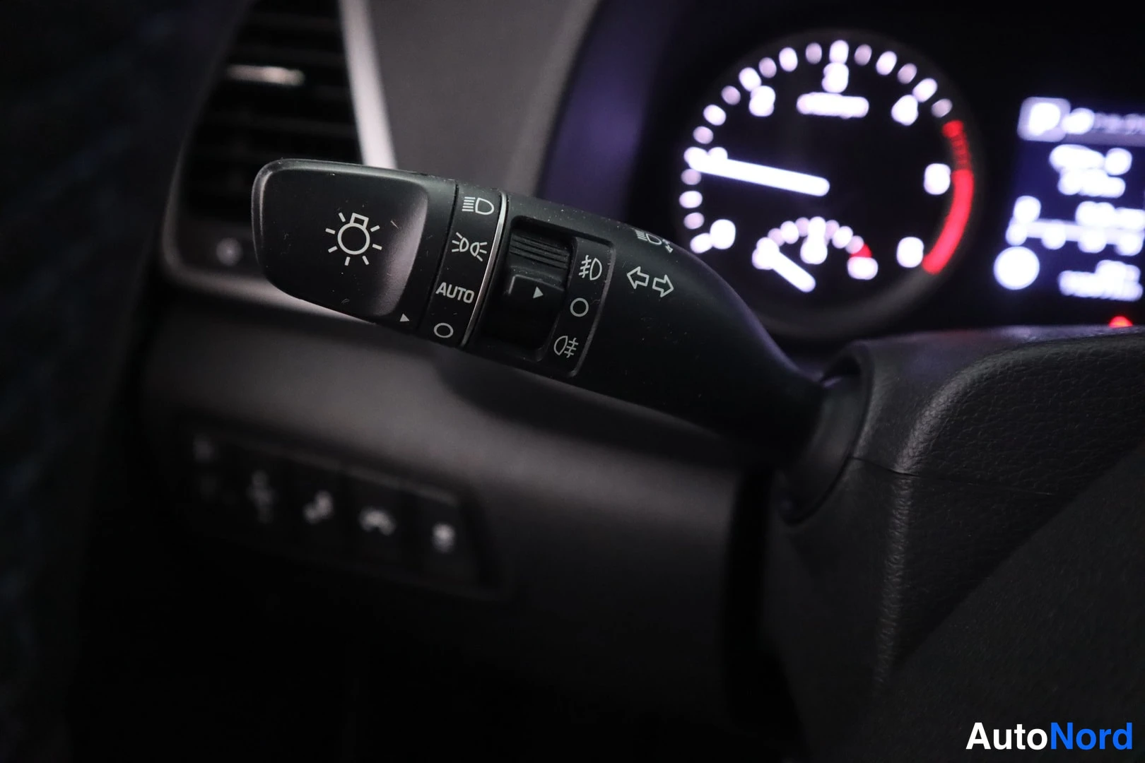 Muu: Valokytkimen varsi AUTO-merkinnällä ja valaistus- ja suuntavilkkusymbolit, takana sumennettu mittaristo.