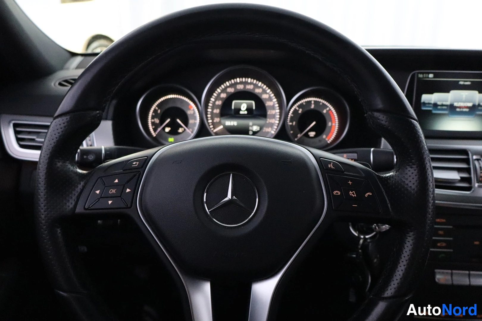 Muu: Mercedes-Benz-automerkki ratti-istuimessa; nahkaratin nappien lisäksi näkyy kojelaudan mittaristo ja keskinäytön näppäimistö.
