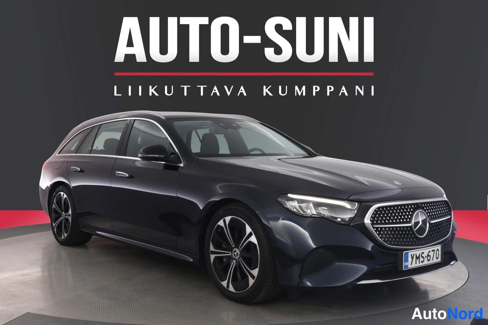 Etuviisto, nokka oikealle: Auton etukulma vasemmalta kuvattuna, tummansininen Mercedes-Benz farmariauto näyttelyalustalla, rekisterikilpi