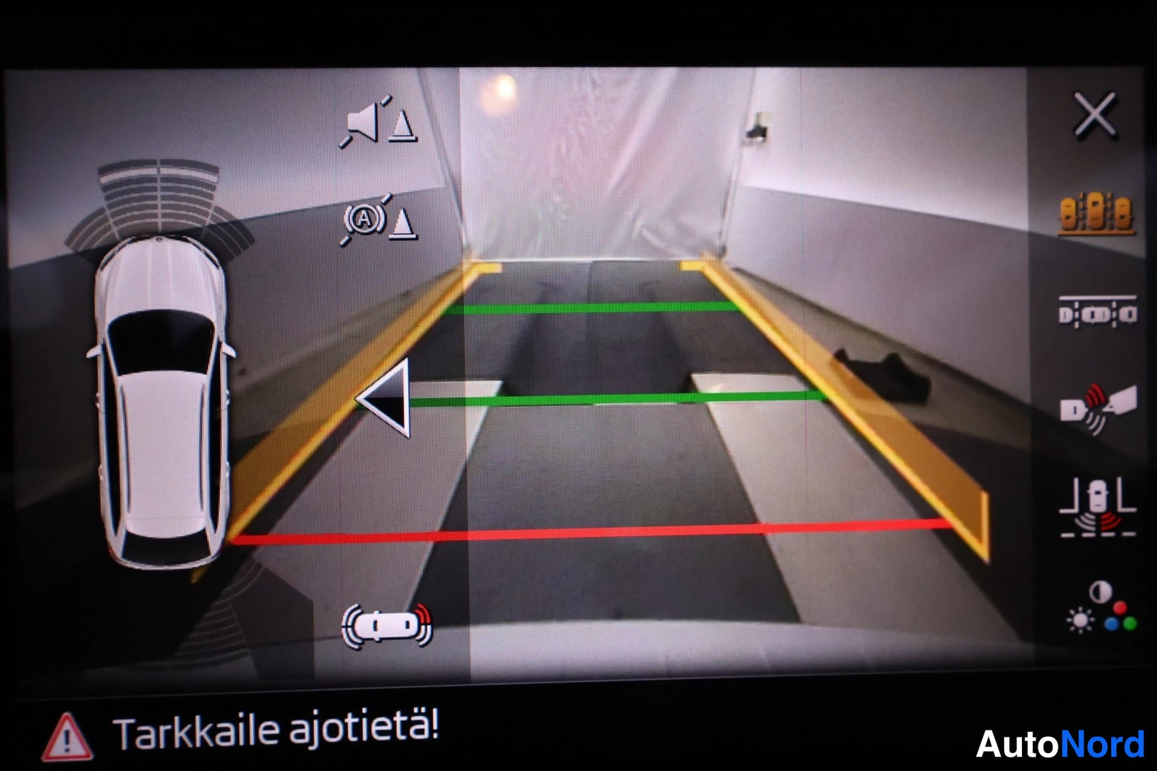 Muu: Infotainment-näytön kuva: ympäristökamerasovellus näyttää auton ylhäältä päin, tiellä vihreät ja punaiset etäisyysviivat sekä varoitustekstin «Tarkkaile ajotiet