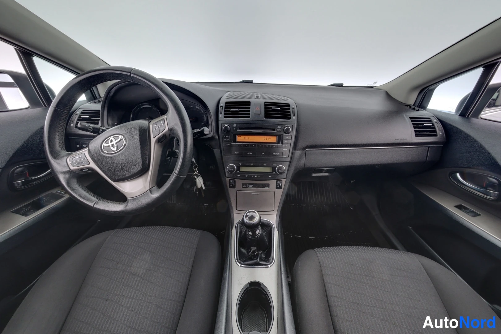 Muu: Toyota Avensisen ohjaamo edestä: Toyota-merkillinen ohjauspyörä, kojelauta ja manuaalivaihtokeppi; kaksi tummaa etuistuinta.