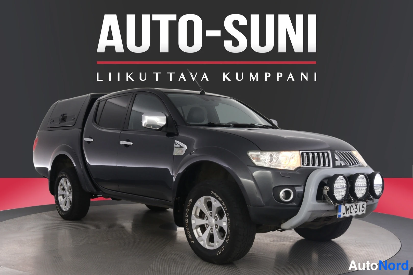 Etuviisto, nokka oikealle: Mitsubishi L200 -mallin tummanharmaa avolava-auto kulmassa edestä oikealta nähtynä, etupuskurissa lisävalot.