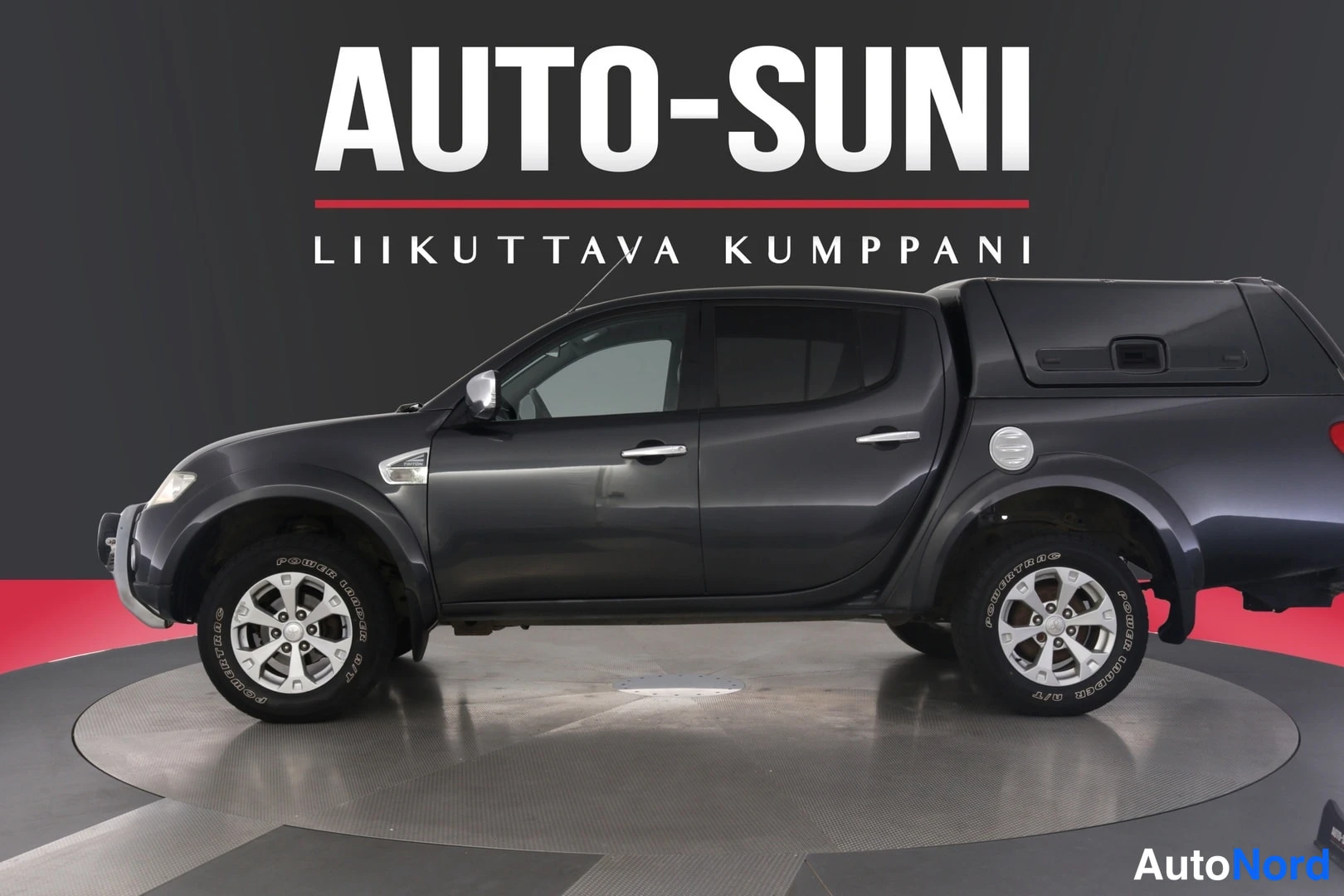 Sivulta: Tummanharmaa Mitsubishin lähikuva autosalon pyörivällä alustalla, kylki ja etuosa näkyvät, taustalla AUTO-SUNI-logo