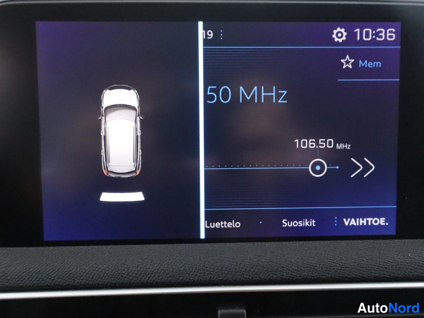 Muu: Infotainment-näyttö jaettuna: vasemmalla auton kuvataustagrafiikka, oikealla radio 106,50 MHz ja valikot Luettelo, Suosikit, VAIHTOE; kuvassa hieman heijastusta