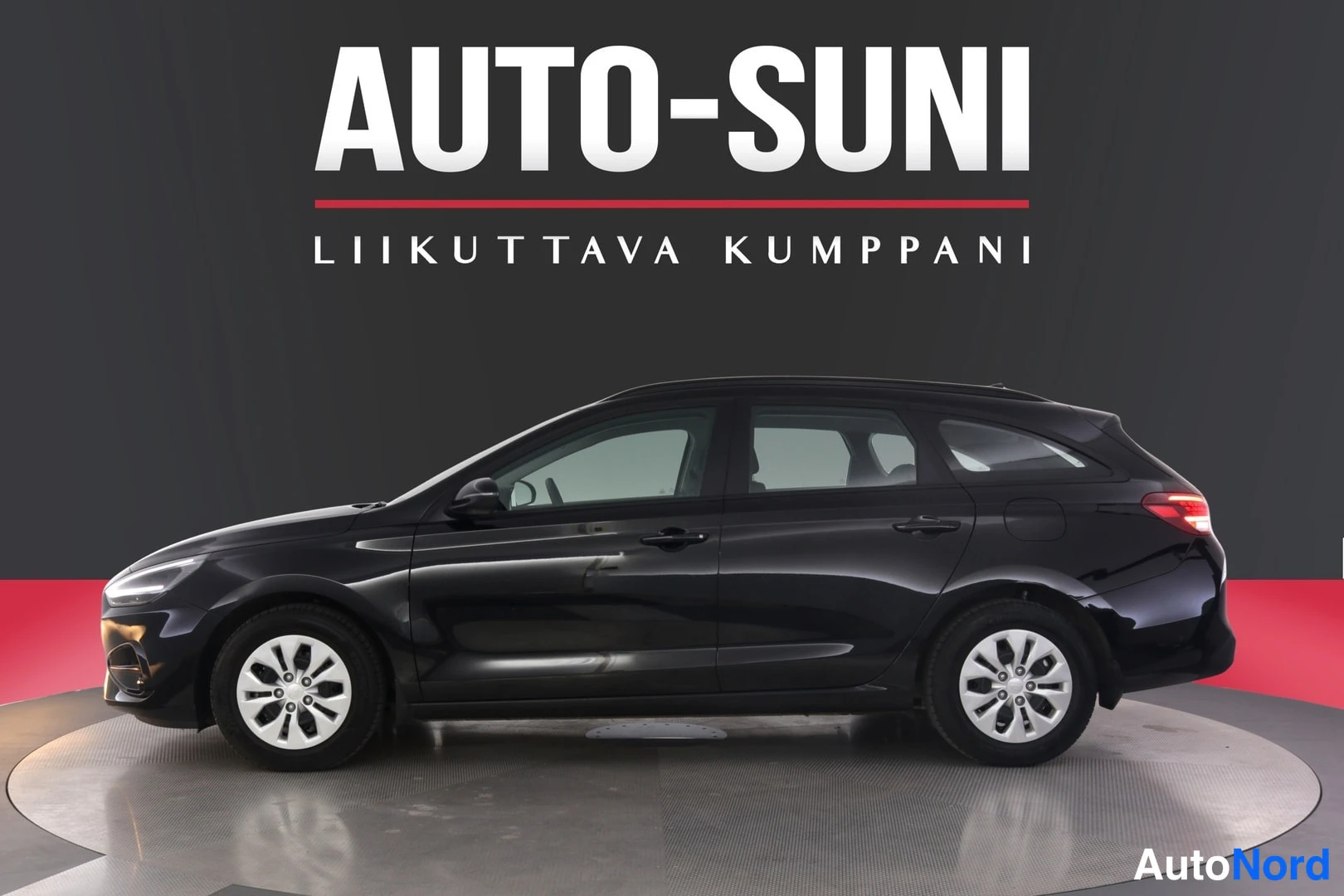 Sivulta: Mustan farmariauto HYUNDAI i30 WAGON 1.6 T-GDI HYBRID COMFORT DCT sivulta kuvattuna, punainen ja tumma studioveto.