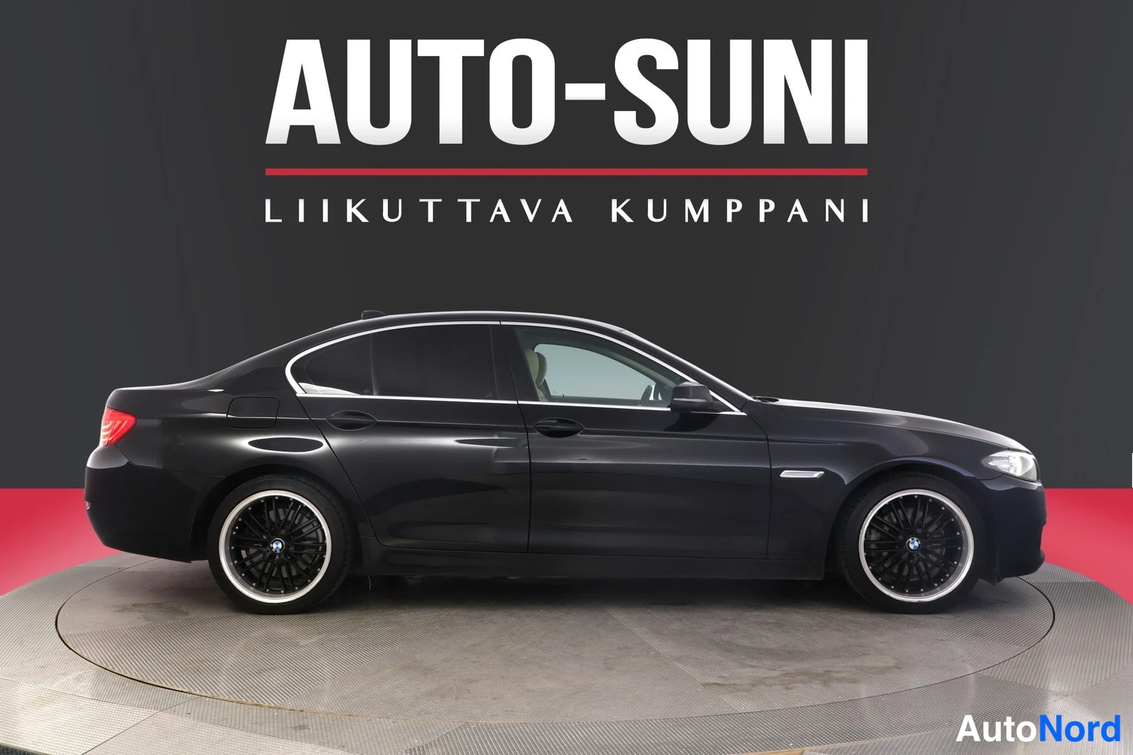 Etuviisto, nokka oikealle: Mustan sedanin sivuprofiili, nokka oikealla, neliväräinen kiinteä metallivanne takana ja edessä, AUTOSUNI-tausta.
