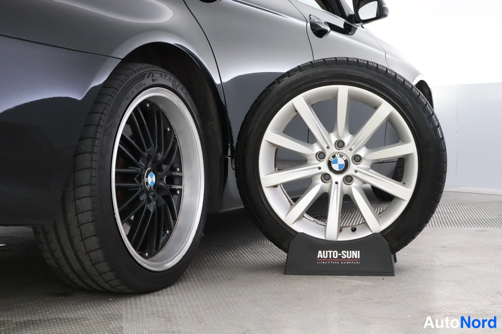 Muu: Auton takapyörän lähikuva tummalla vanteella; vieressä sijaisvanteena BMW-merkitty hopeinen 10-puolainen vanne AUTO-SUNI-telineessä.