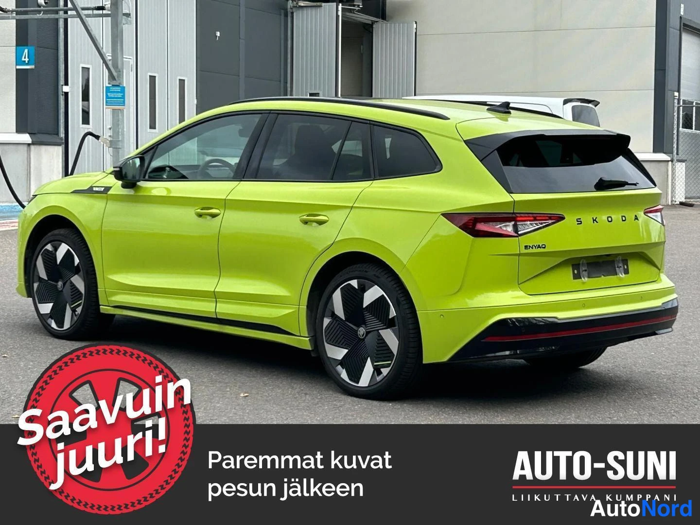 Takaviisto: Passenger-side rear view of the car parked near a building. The flanks (doors) and the rear tailgate with 'SKODA ENYAQ' badging are both visible. The camera pos