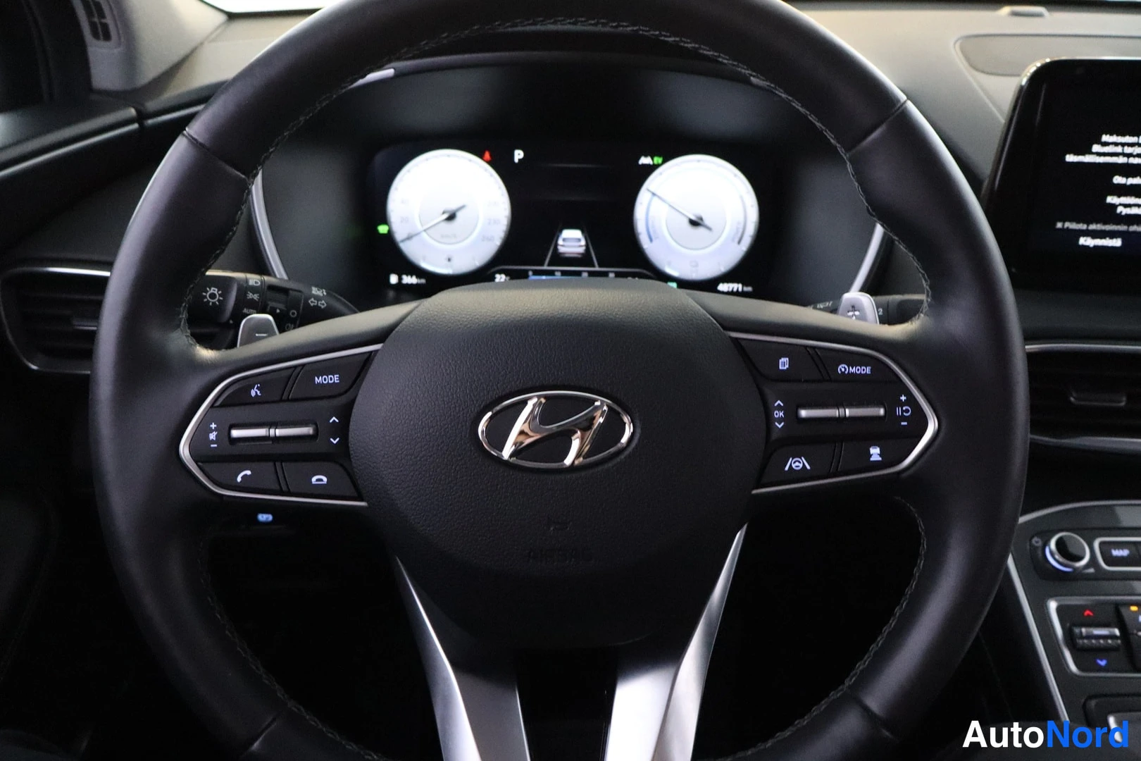 Ohjaamo: Ohjaustahdin keskeinen Hyundai-logo, ympäröivät ohjaimet ja takana oleva digitaalinen kokopainike. Ohjaustahdin molemmin puolin näkyvät äänenvoimakkuuden säädin