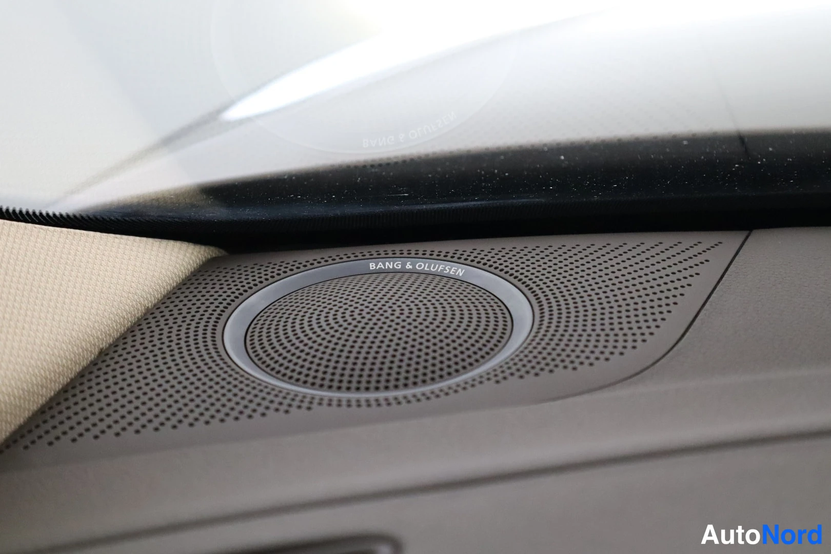 Muu: Kojelaudan Bang & Olufsen -merkitty kaiutin pölyn suodattimessa; teksti BANG & OLUFSEN kaiuttimen kehällä, beige verhoilu vasemmalla.