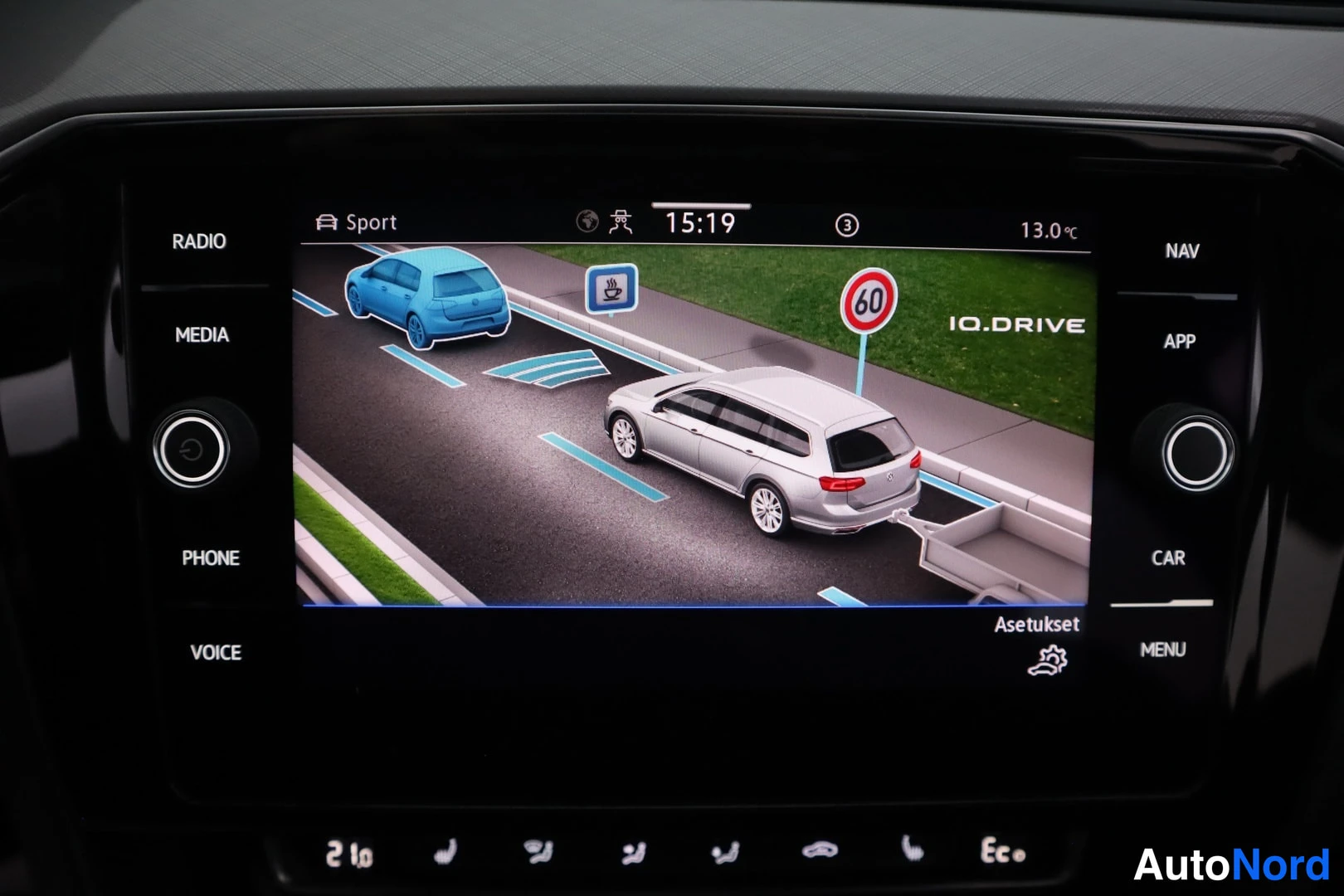 Muu: Infotainment-näyttö näyttää IQ.DRIVE-avustajan animaation, jossa hopea farmariauto seuraa sinistä autoa ja tien laidalla on 60-merkki.