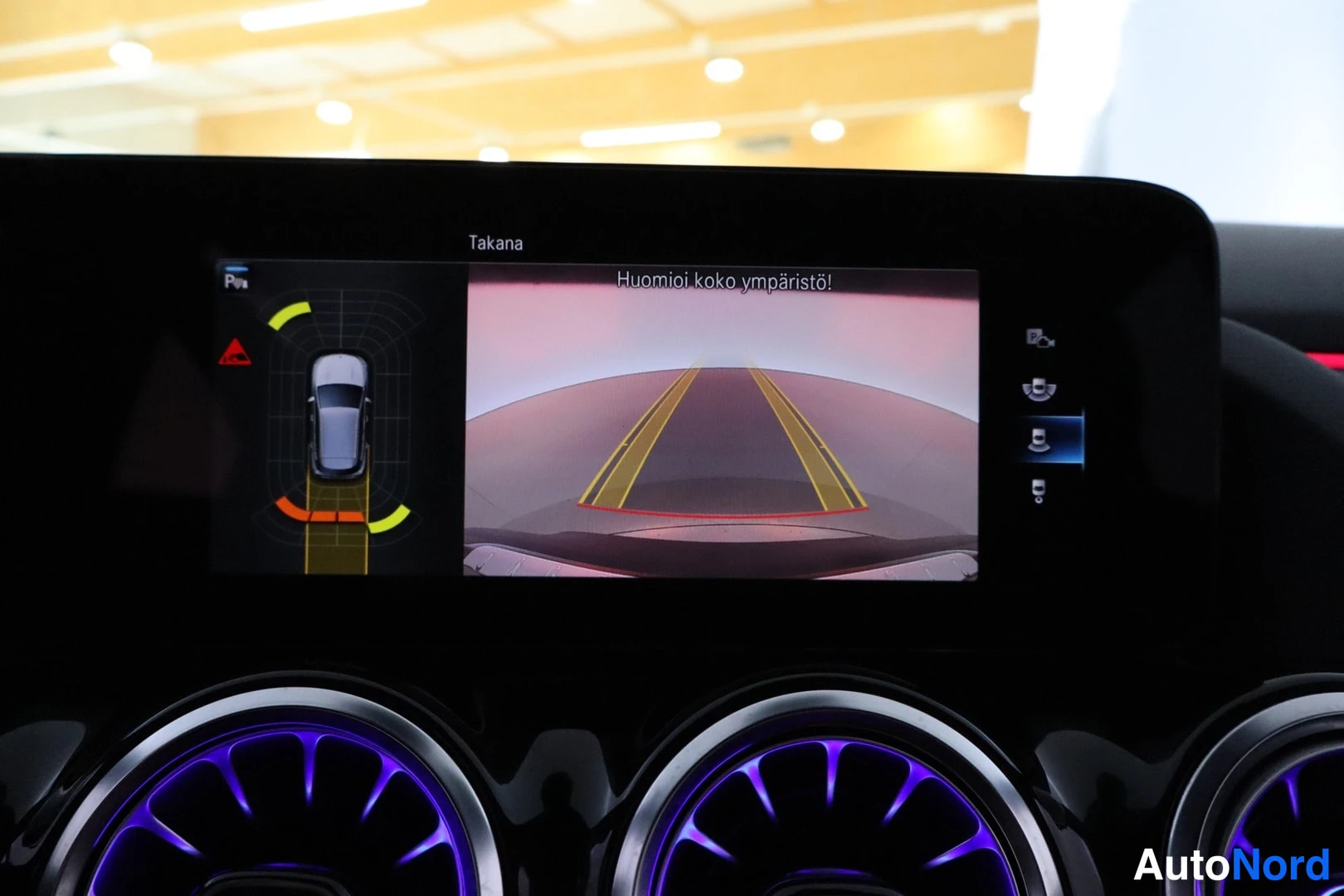 Muu: Infotainment-näyttö näyttää peruutuskameran kuvan ja huomion 'Huomioi koko ympäristö!', alla tunnelmavalaistut ilmastointisuuttimet.