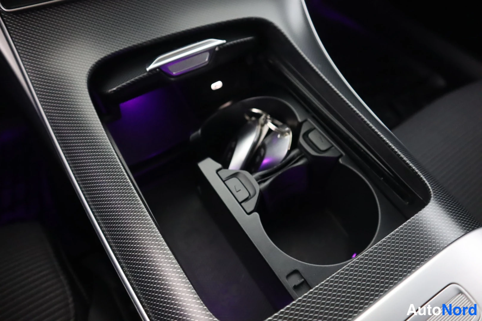Yksityiskohta: Close-up of the center console storage area containing cup holders and the electronic gear selector, illuminated by purple ambient lighting.