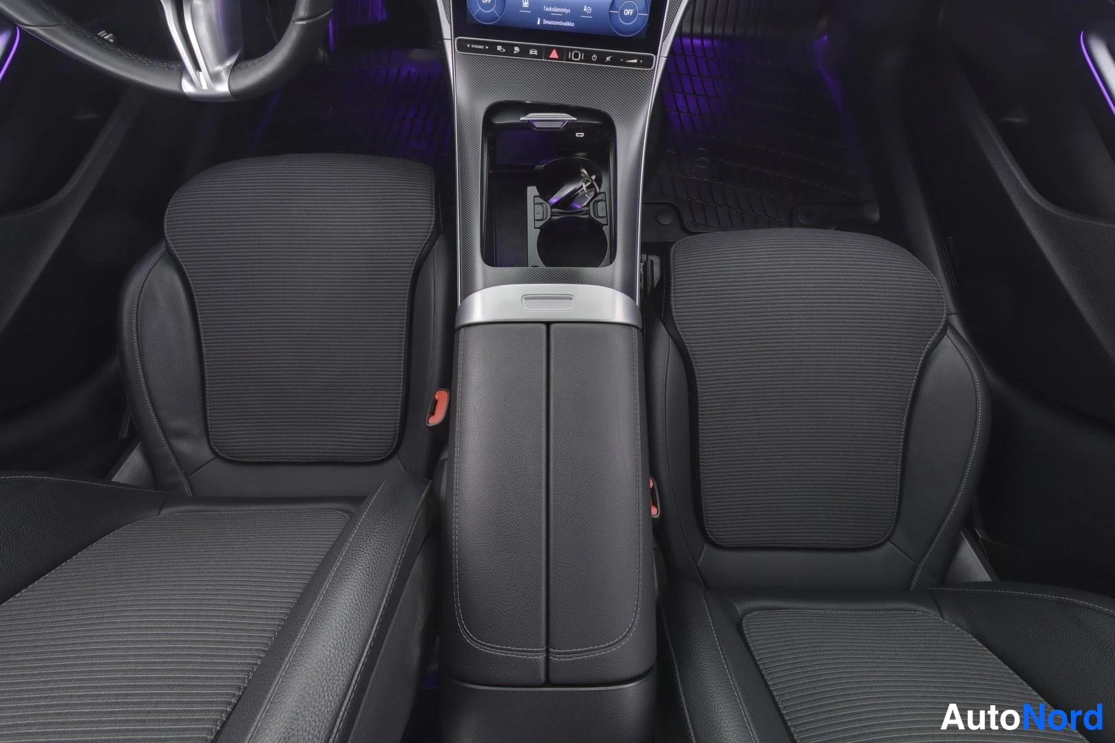 Etuistuimet: Interior view of the front cabin showing two black leather seats with fabric inserts, a center console between them, and part of the steering wheel visible on t