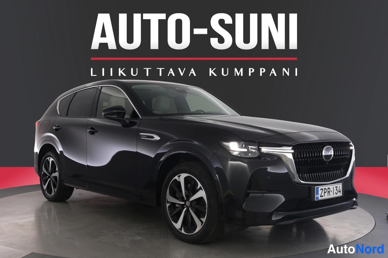 Etuviisto, nokka oikealle: Musta Mazda CX-60 -maastokuva kolmesta neljäsosasta etupää oikealla, rekisteritunnus, myyntihallissa tummaa taustaa vasten.