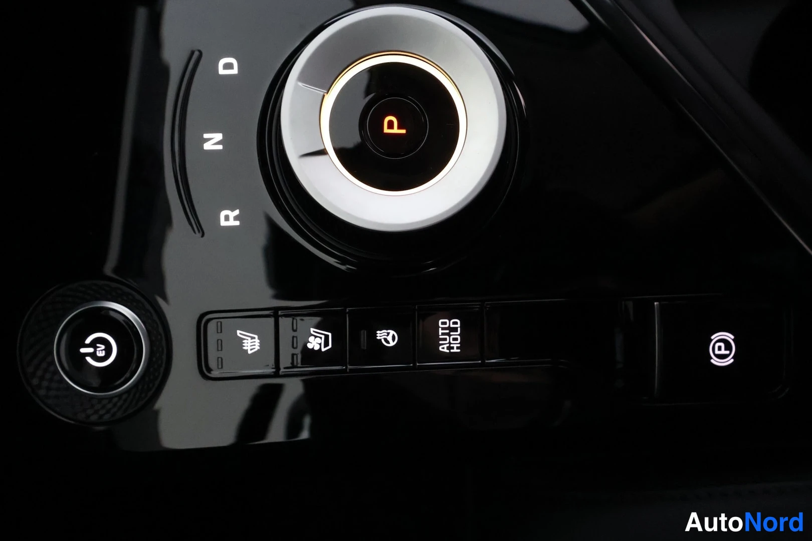 Yksityiskohta: Close-up of the rotary automatic gear selector in Park position with the brake pedal release button and parking brake button nearby.