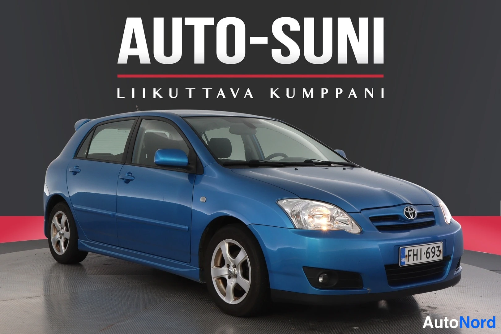 Etuviisto, nokka oikealle: Blue Toyota Corolla five-door hatchback photographed from a front three-quarter angle with the nose pointing left against a dark grey background.