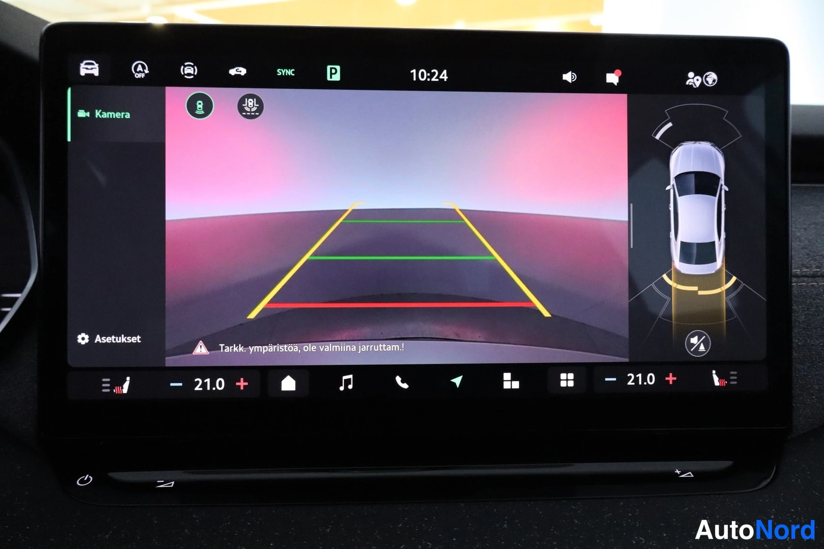 Muu: Auton infotainment-näyttö näyttää peruutuskameran kuvan parkkirutilla ja tekstillä 'Tarkk. ympäristöä, ole valmiina jarruttamaan!'.