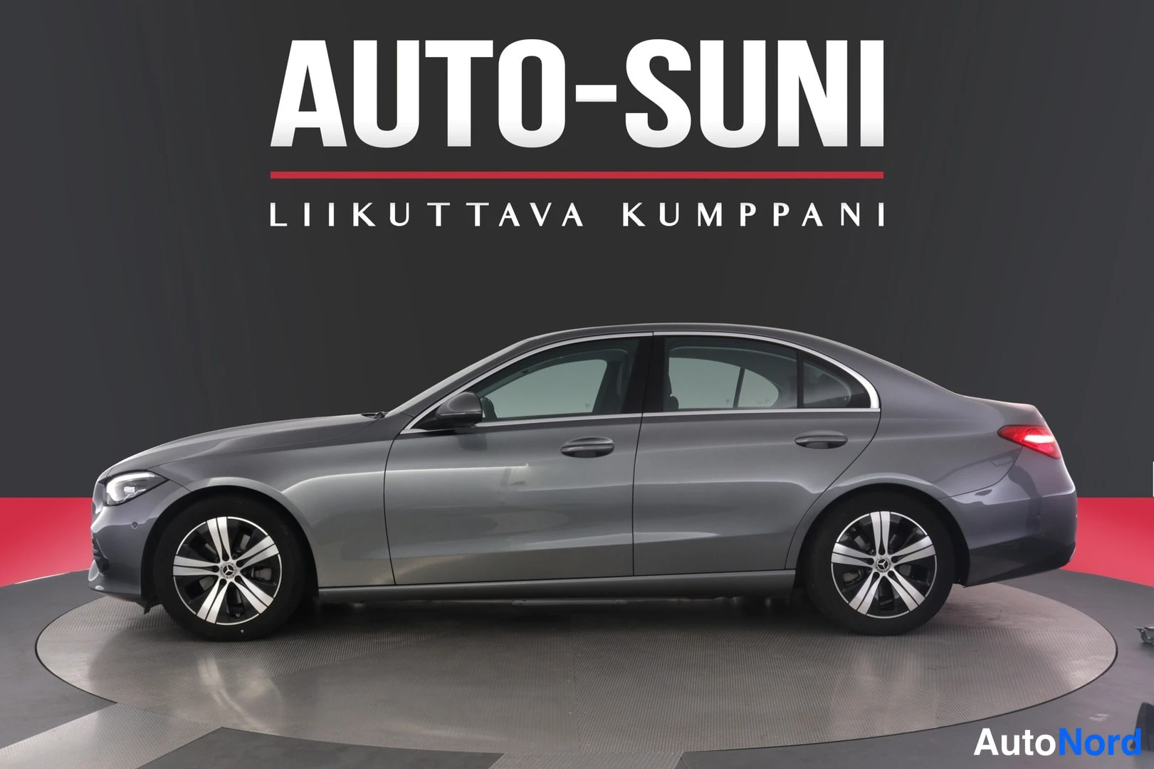 Sivulta: Auto-Suni-logoa yläosassa; harmaa Mercedes-Benz-sedan täydessä sivuprofiilissa vasemmalle etunohtuna kääntyneellä lavalla