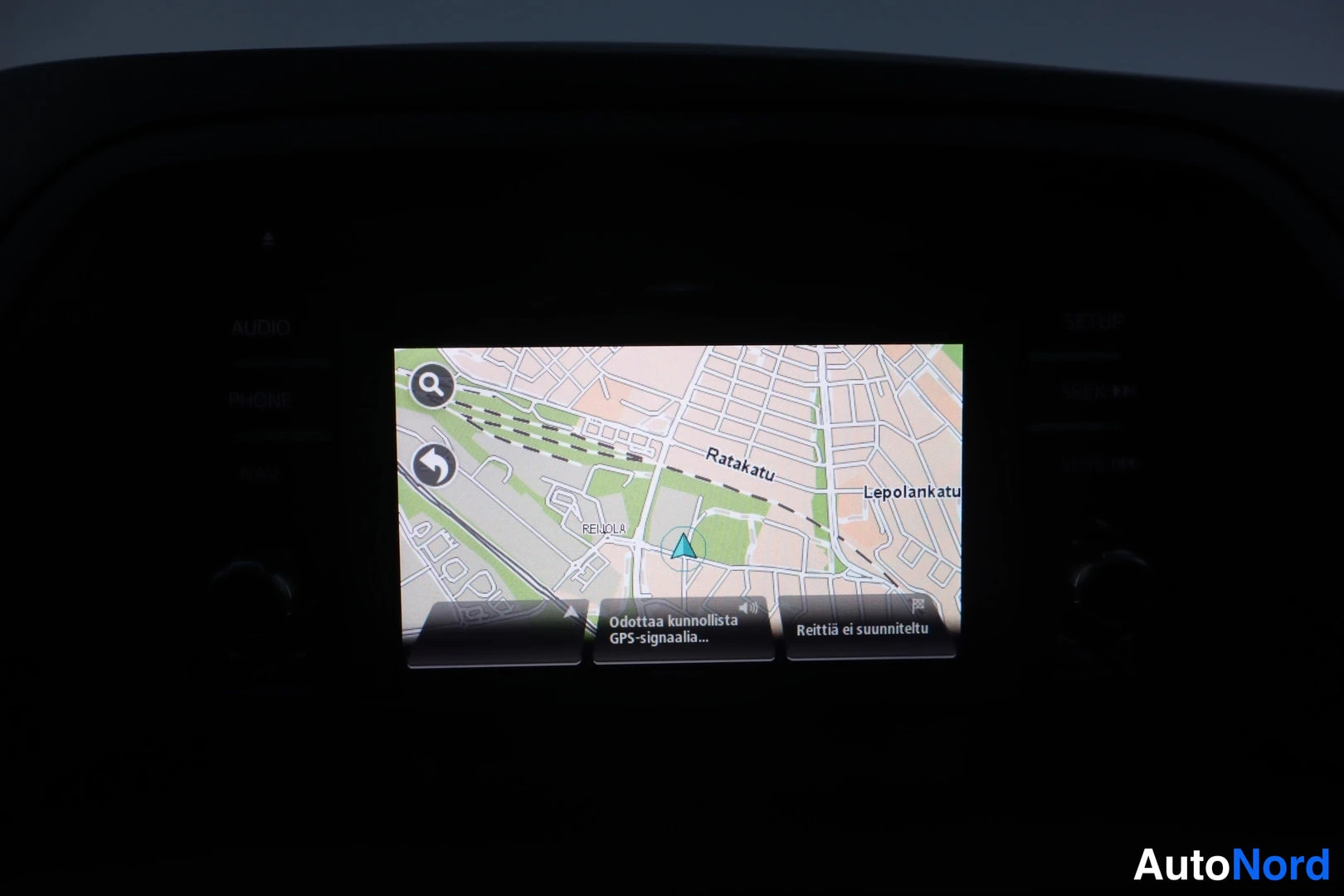 Yksityiskohta: Tummassa ohjaamossa infotainment-näyttö näyttää navigointikarttaa (Ratakatu, Lepolankatu) ja tekstiä 'Odottaa kunnollista GPS-signaalia...', 'Reittiä ei suunnit