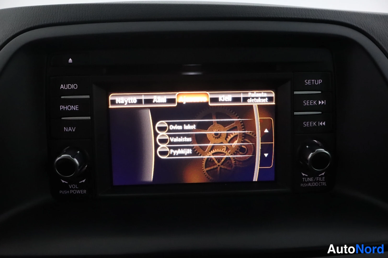 Yksityiskohta: Infotainment-näytön asetusvalikko välilehdillä Näyttö, Ääni, Ajoneuvo, Kello ja Palauta oletusasetukset; valinnat Ovien lukot, Valaistus, Pyyhkäijät. Reunapaini