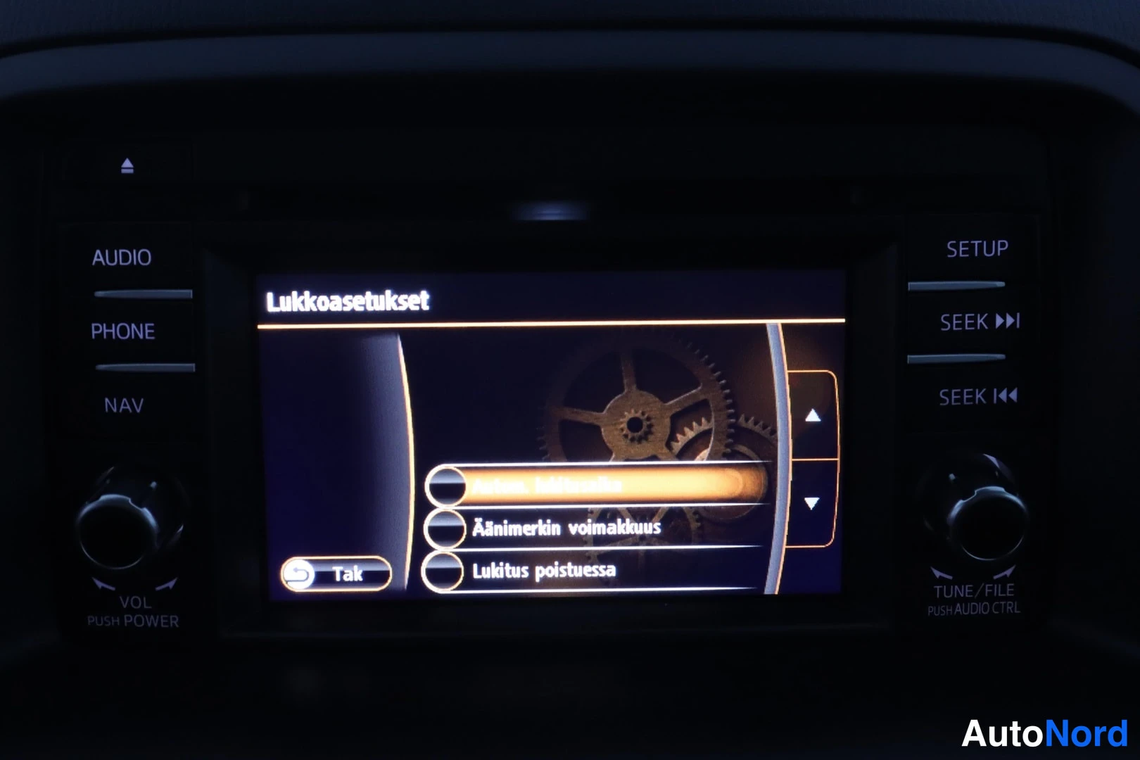 Yksityiskohta: Tummassa ohjaamossa infotainment-näytön 'Lukkoasetukset'-valikko vaihtoehdoilla Äänimerkin voimakkuus ja Lukitus poistuessa sekä Tak-painike; AUDIO-, PHONE-, NA