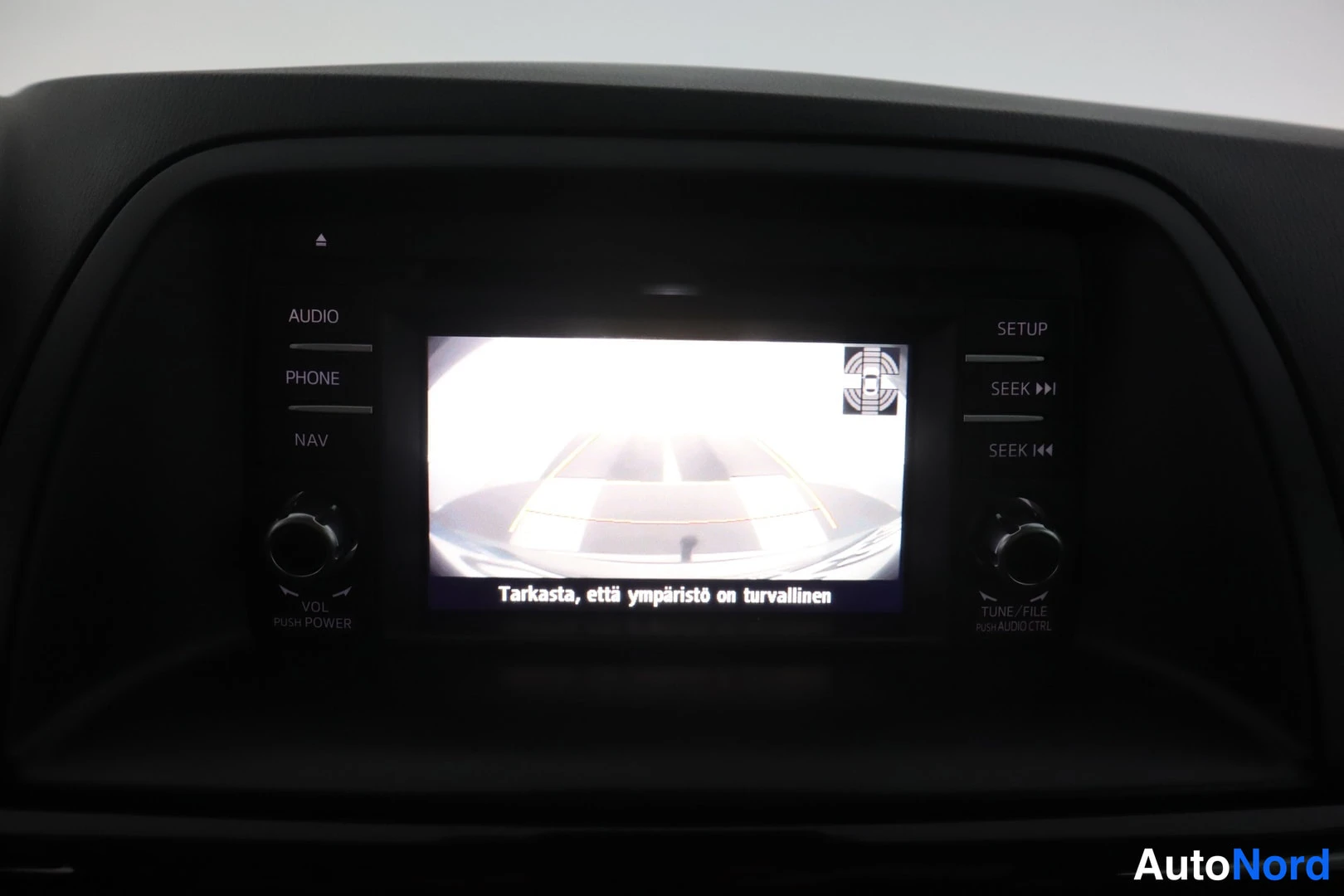 Muu: Kojelaudan infotainment-näyttö näyttää peruutuskameran kuvan; tekstinä Tarkasta, että ympäristö on turvallinen.