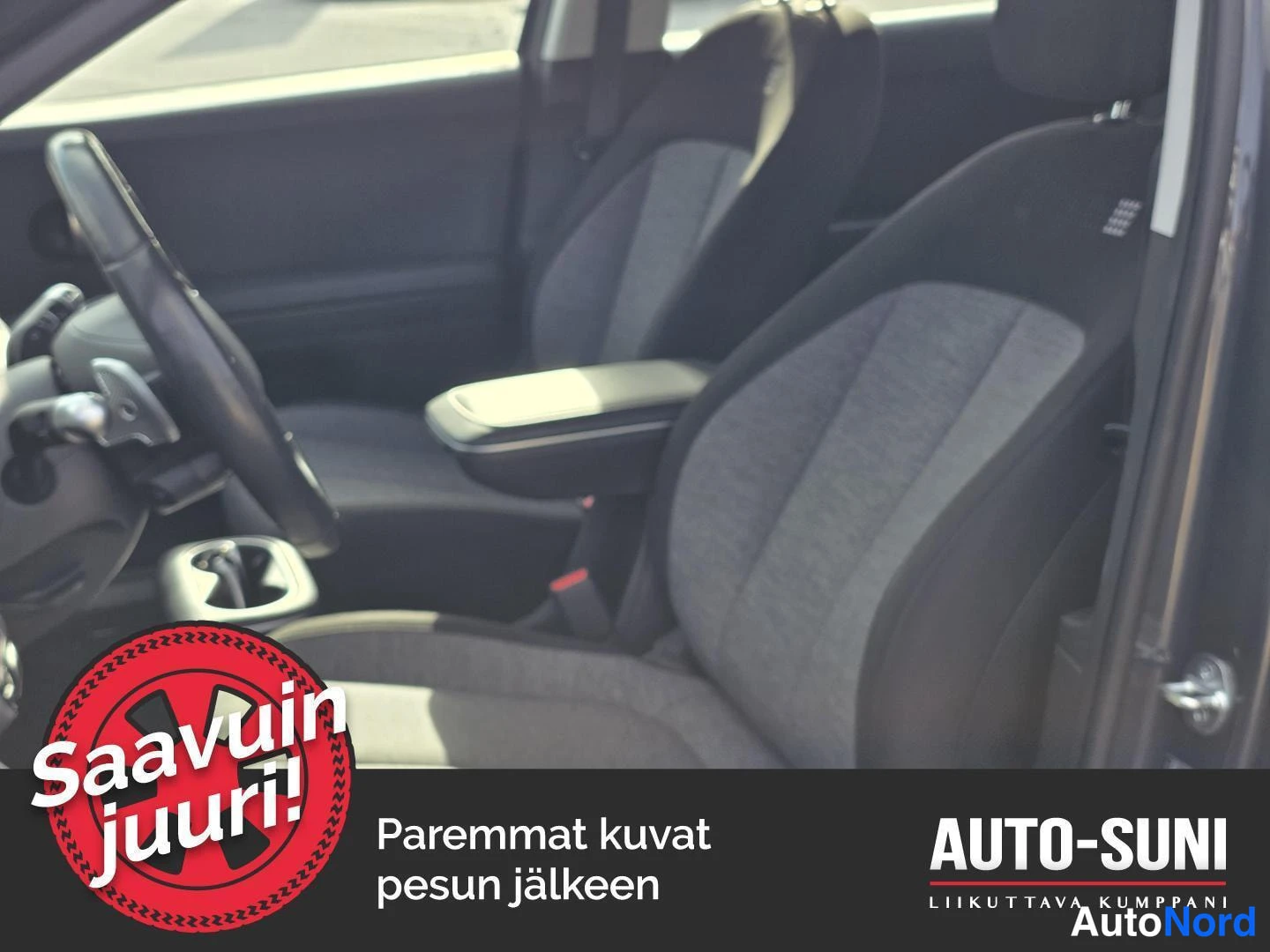 Muu: Auton etupenkit ja kojelauta sisätiloista kuvattuna; kankaiset istuimet, ohjauspyörä ja keskusvarsi näkyvät. Kuvassa mainosgrafiikka: 'Saavuin juuri!' sekä teks