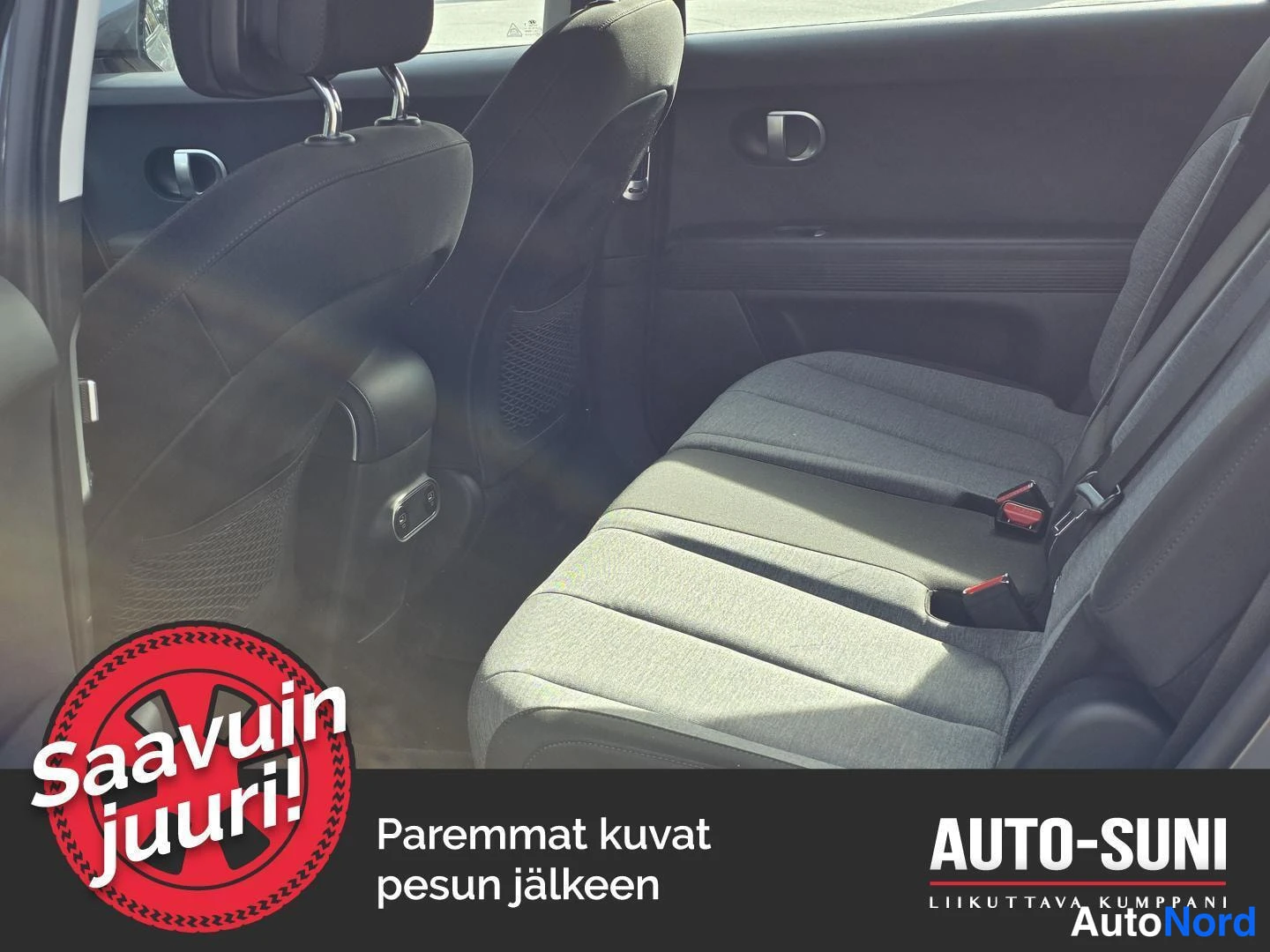 Muu: Auton takaistuin sisätiloista kuvattuna; kangaspäällysteinen penkki, takana istuinten takapinnat ja ovi näkyvät. Kuvassa mainosgrafiikka: 'Saavuin juuri!', teks