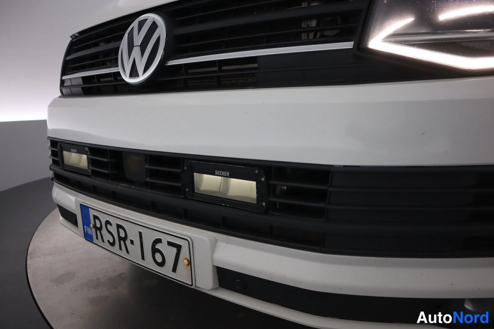 Edestä: Etuvaihto: valkoisen Volkswagenin etusäleikkö VW-emblemillä, pohjassa kaksi SEEKER-tyyppistä lisävaloa ja rekisterikilpi.