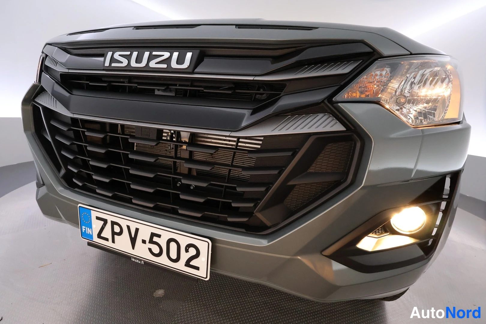 Edestä: Ajetun auton etupää lähikuvassa: ISUZU-merkki jäähdyttäjäsuojukessa, sumuvalo päällä, rekisterikilpi.