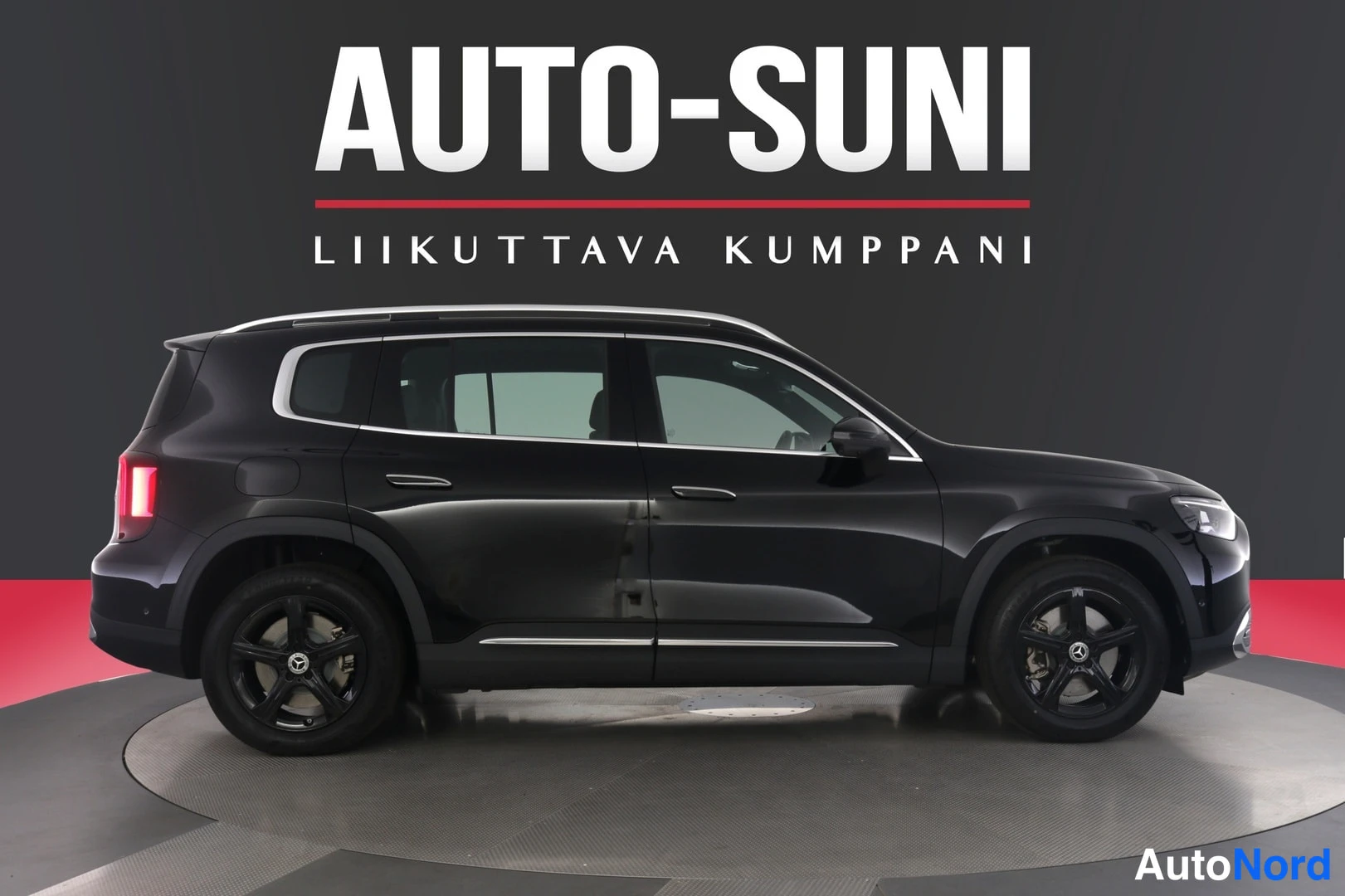 Etuviisto, nokka oikealle: Musta Mercedes-Benz GLB -maasturi sivusta oikealta katsottuna, kuvassa oikean puolen ovet ja pyörät, taustalla tumma seinä ja punainen kaista.